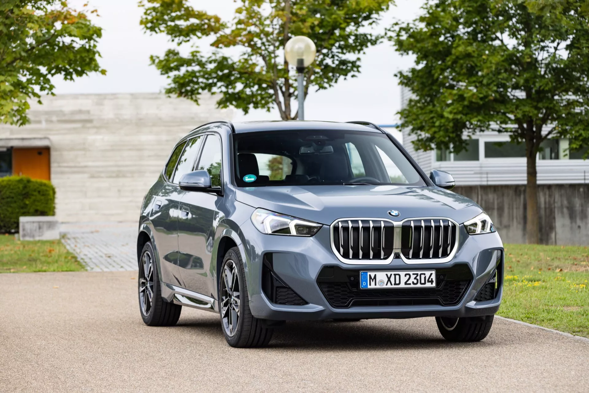 Lyxig crossover SUV BMW X1 (U11) 2022 i mörkgrå metallic, med LED-strålkastare och aerodynamisk frontstötfångare, parkerad i en landskapsarkitekturerad stadspark