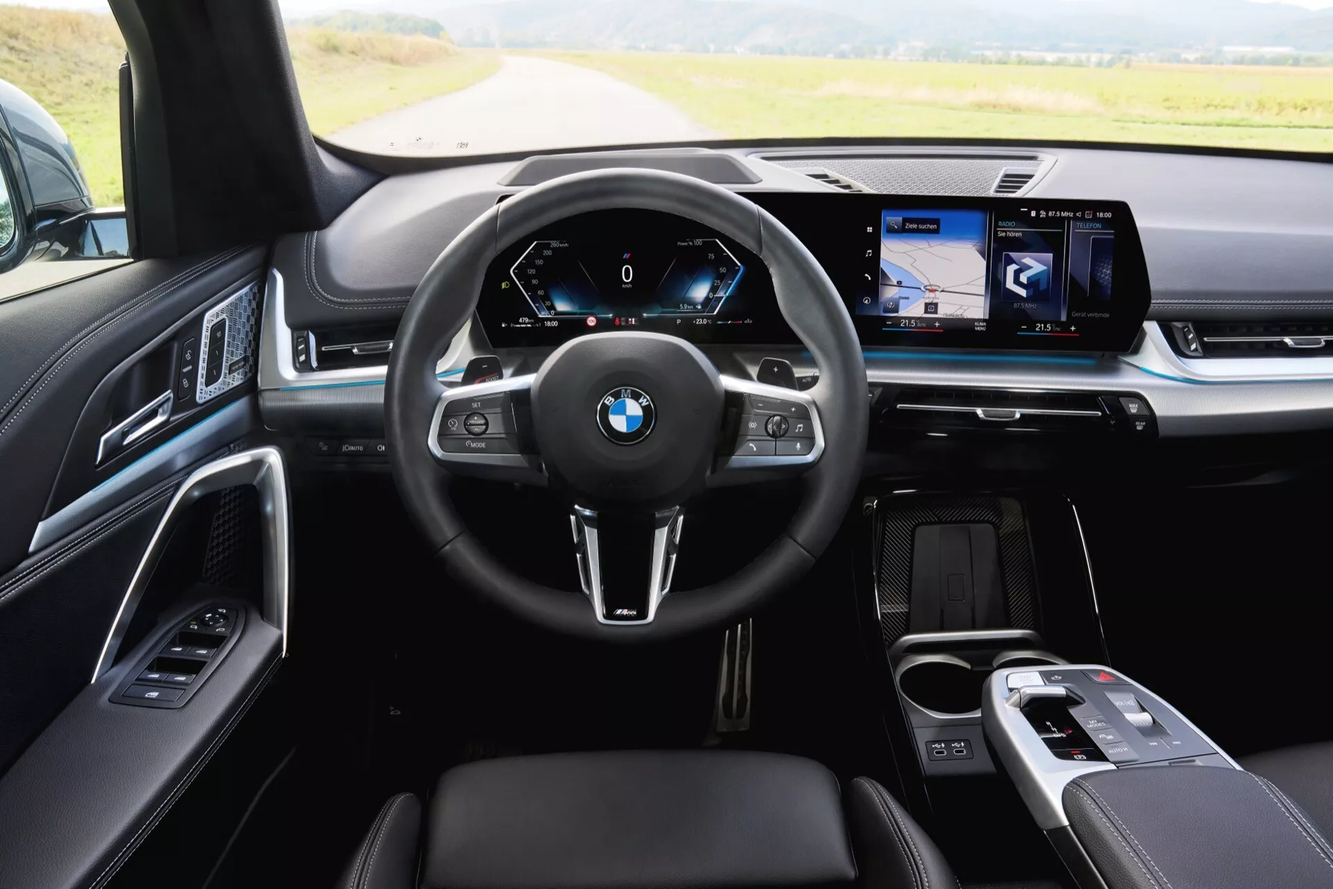 Interiörvy av BMW X1 (U11) 2022 från baksätet som visar förarens kontrollzon med multifunktionsläderratt, digital instrumentpanel och widescreen navigeringssystem på instrumentbrädan