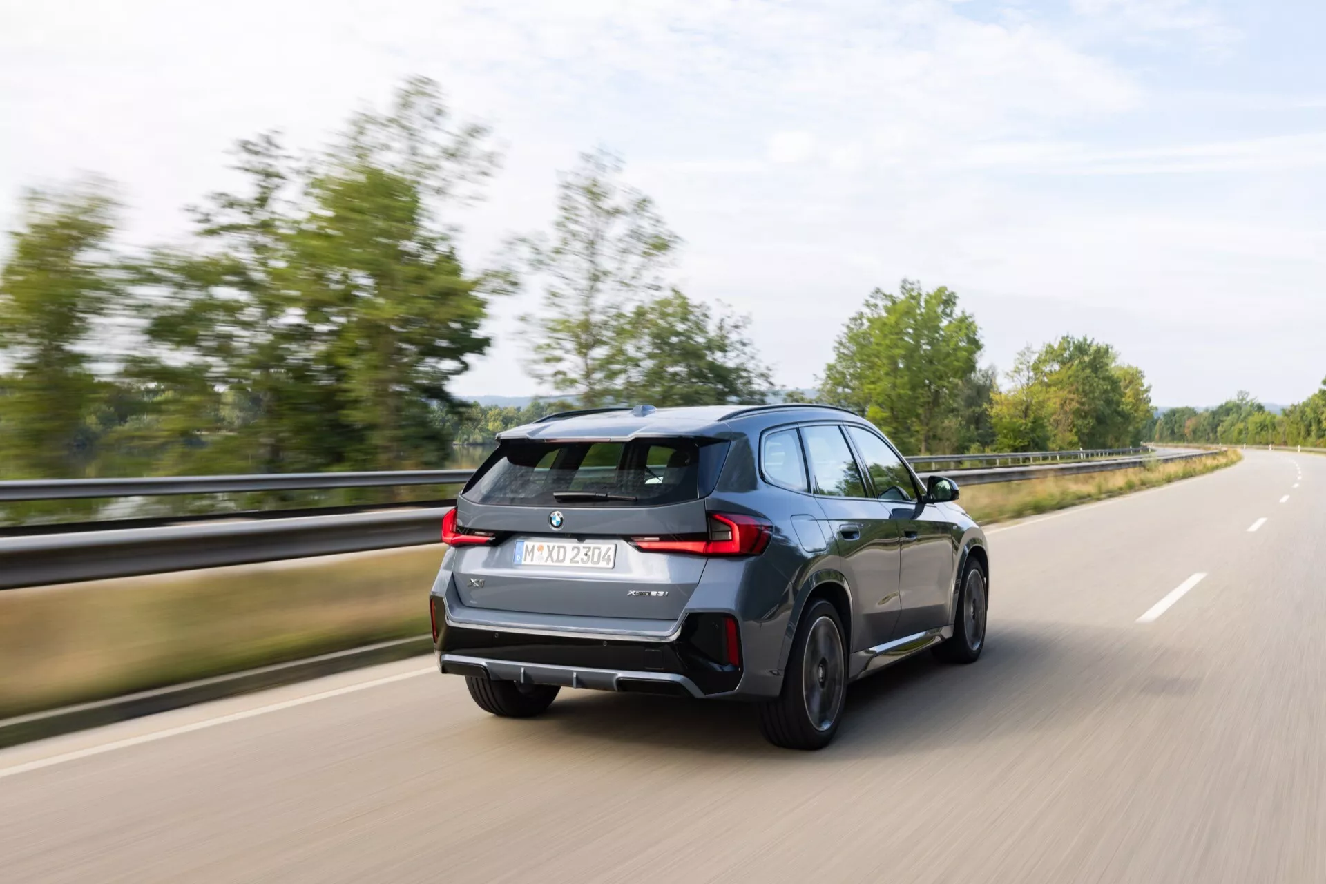 Dynamisk trekvarts bakre vy av BMW X1 (U11) 2022 i rörelse på en väg, framhävande LED-bakljus, bakre diffusor, sportig design och suddiga hjul som indikerar hastighet