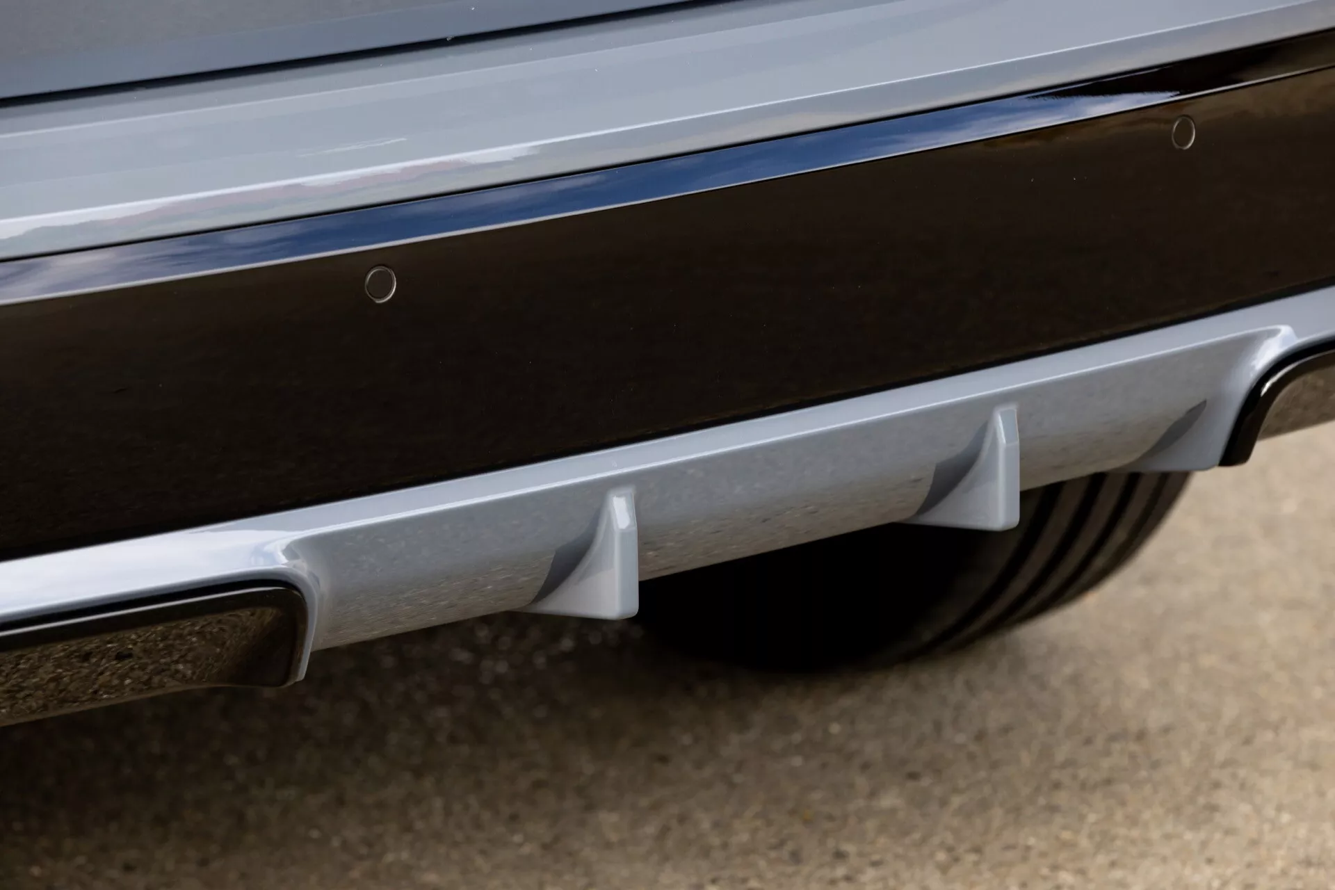 Närbild i lågt vinkel av bakre stötfångaren på BMW X1 2022 (U11) som framhäver egenskaper inklusive en bakre diffusor med vertikala fenor, ett kromat avgasrör, parkeringsassistanssensorer, reflekterande element och kromad trim, vid sidan om en vy av ett sportigt flerspakat hjul och däck.