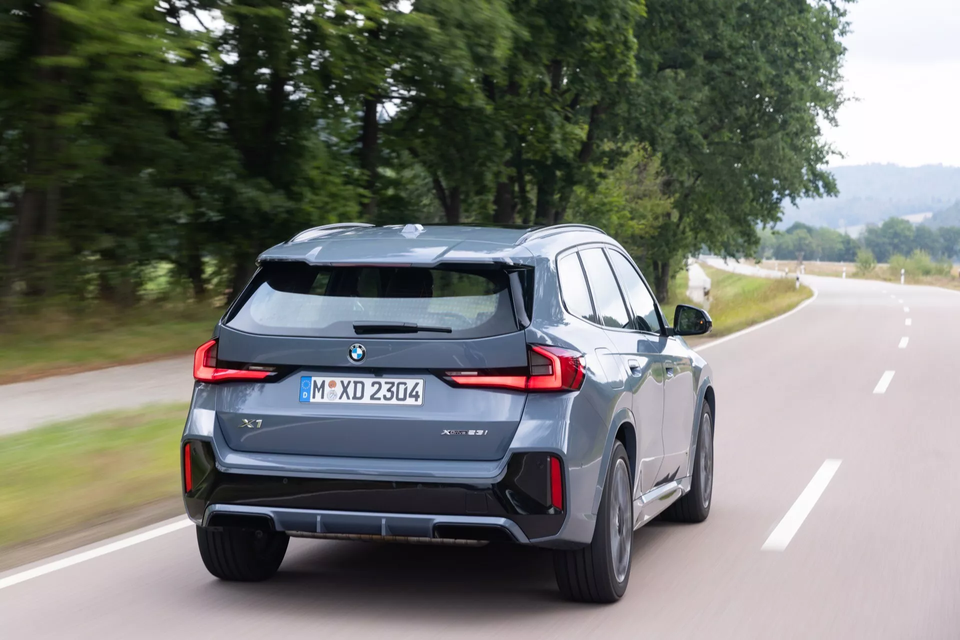 BMW X1 (U11) 2022 i metallisk färg med distinkta bakljus och dubbla avgasrör, visar bak- och delvis höger sida vy i rörelse med suddig grön bakgrund, möjligen indikerande avancerade funktioner som adaptiv belysning och hybrid drivlina