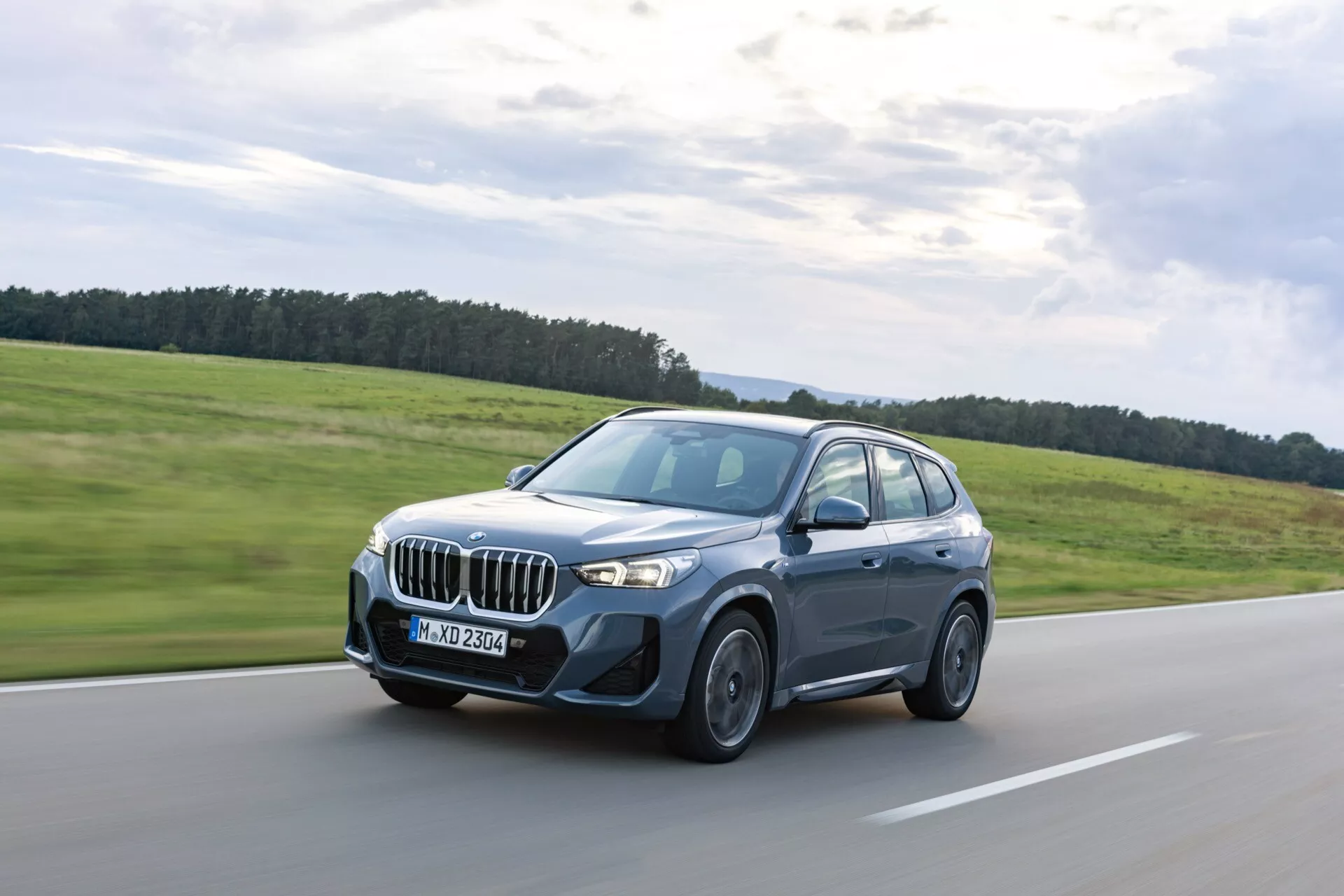 Bilden visar en BMW X1 (U11) SUV från 2022 i rörelse på en väl underhållen väg. Bilden, tagen från vänster främre sida, visar den karakteristiska BMW-njurgrillen, eleganta LED-strålkastare, aerodynamisk främre stötfångare och dynamisk design. Fordonet är i en mörkblå eller grafitton med svarta hjul och har lätt tonade fönster, och rör sig diagonalt mot vänster. Suddiga hjul betonar dess rörelse, medan subtila designdetaljer antyder avancerade funktioner som adaptiv belysning och premium legeringshjul.