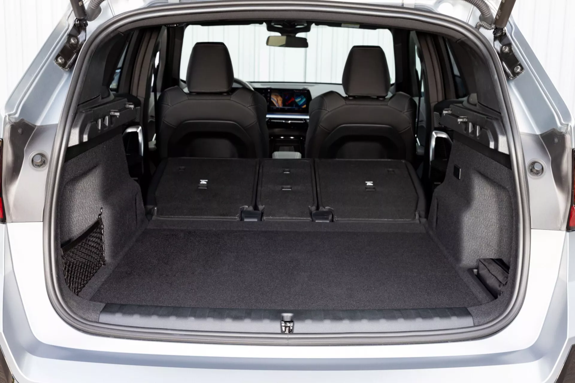 Interiörvy av BMW X1 (U11) 2022 som framhäver lastutrymmet med platt lastgolv och baksäten fällda i 60/40-split, med ett nätat förvaringsfack, sidoförvaringsfack, baksätes mugghållare och en glimt av pekskärmsinfotainmentsystemet från bakre perspektivet