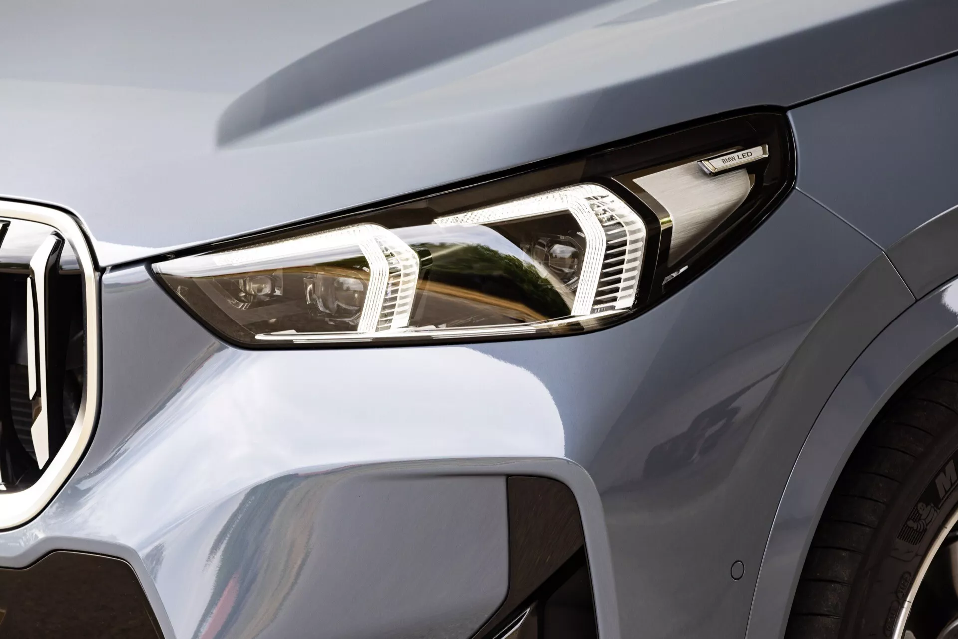 Närbild av den främre vänstra delen av en BMW X1 2022 (U11), med fokus främst på den moderna LED-strålkastaren med tydliga ljuselement och dagsljus (DRL). Motorhuvens framkant syns i det övre högra hörnet och lutar ner mot ett delvis synligt främre galler på vänster sida. Den metalliska eller pärlemorsfärgade lacken speglar en slät, glansig finish som framhäver de skulpterade linjerna och de aerodynamiska egenskaperna hos bilens design. Fotot är taget från en låg till medelhög vinkel och fångar detaljerade aspekter av strålkastaraggregatet, framstöten och gallret.