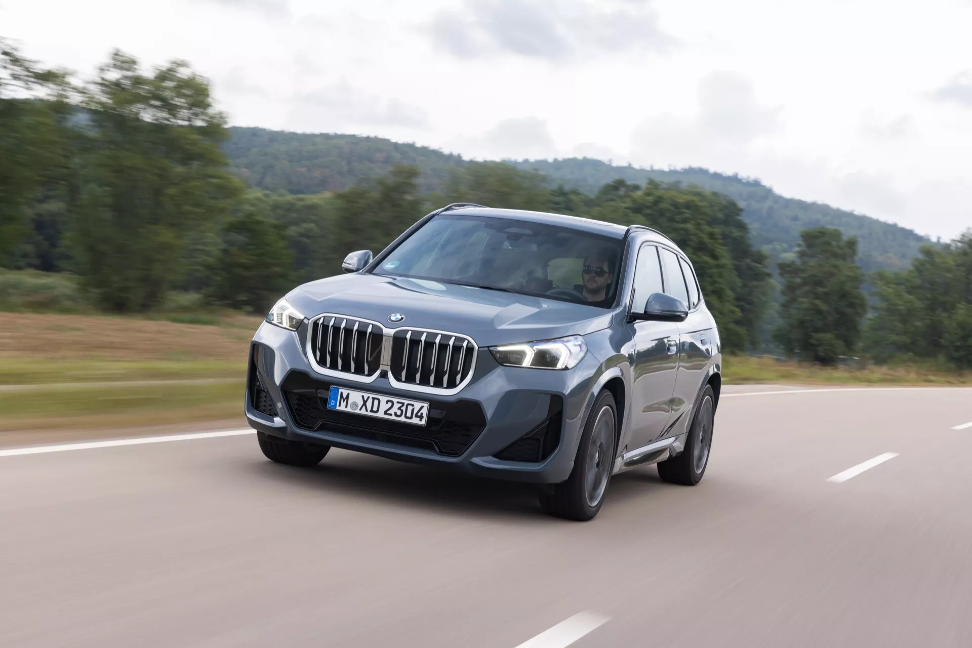 BMW X1 (U11) 2022 i rörelse med fokus på dess distinkta njurformade grill, vinklade LED-strålkastare och lyxiga lättmetallfälgar, som visar avancerade funktioner som adaptiv belysning, parkeringssensorer och möjligt surround-kamerasystem, mot en landsbygdsbakgrund