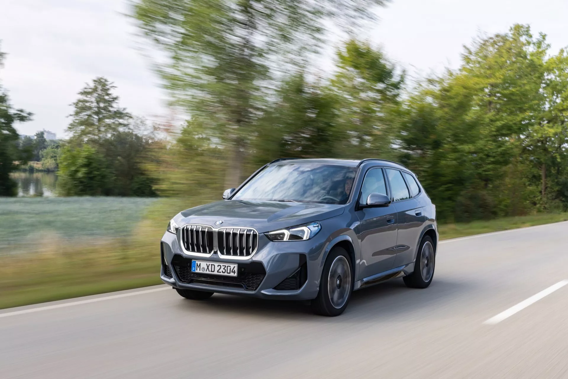 Lyxig SUV BMW X1 (U11) 2022 i rörelse på en offentlig väg omgiven av vegetation på dagen, visar sin design och teknik från en framåtvänd trekvarts vinkel med lätt rörelseoskärpa
