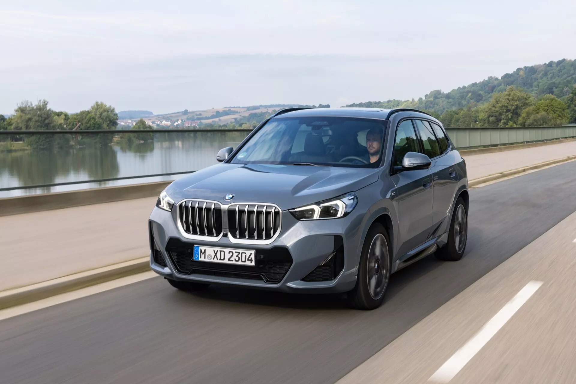 2022 BMW X1 (U11) lyxig crossover SUV i rörelse på en slät väg vid en vattenkropp, som visar dynamisk design, LED-strålkastare och avancerade teknikfunktioner