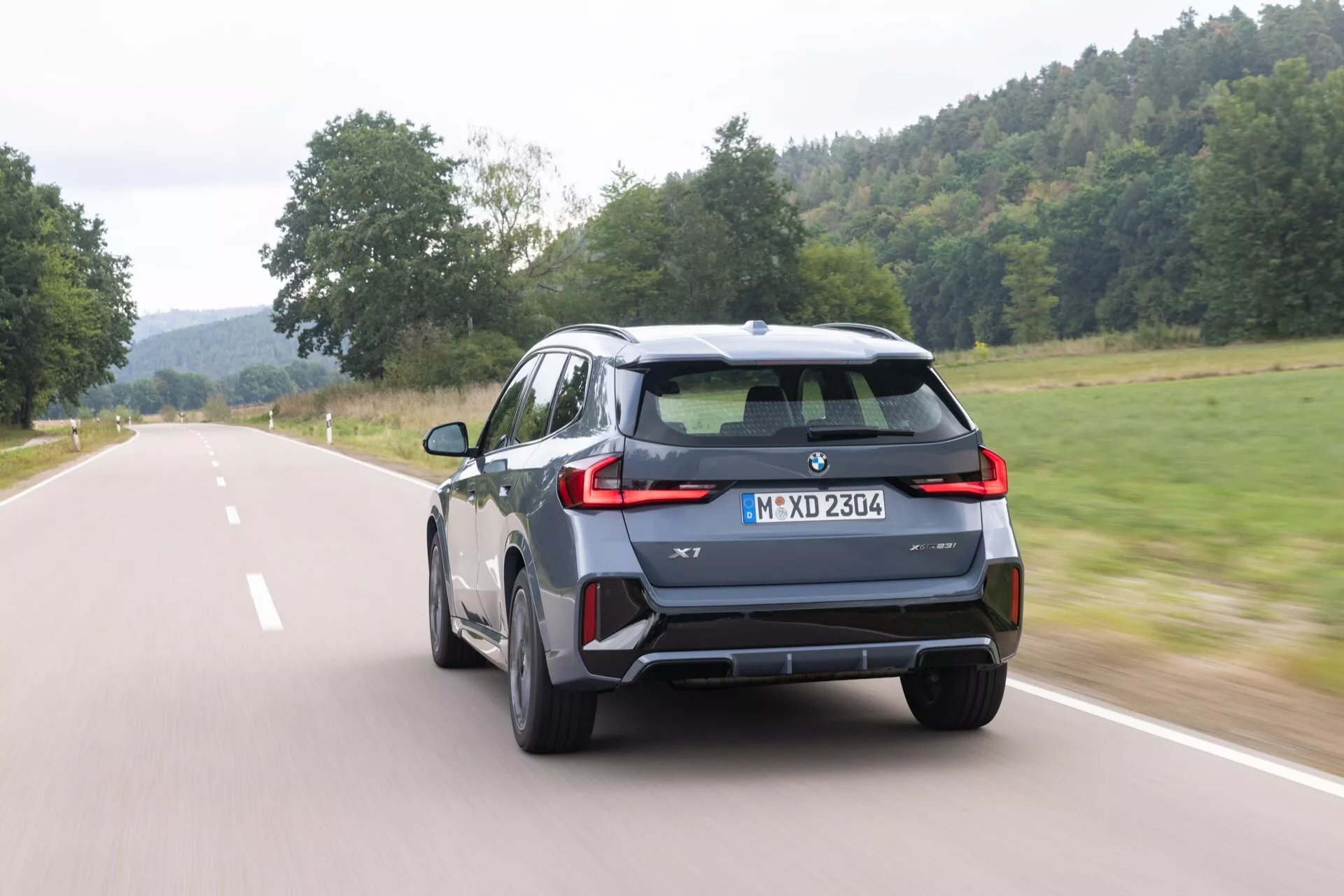 Dynamisk vy bakifrån av en BMW X1 2022 (U11) SUV som kör på en landsväg, som visar moderna LED-baklyktor, potentiella parkeringsassistanssensorer eller reflexer på den bakre stötfångaren, centralt monterad utrymme för registreringsskylt och backkamera, hajfensantenn på taket, stilfulla lättmetallfälgar, integrerad bakspoiler och märkning som indikerar modell eller trimnivå, allt mot en lugn lantlig bakgrund under molnigt dagsljus.