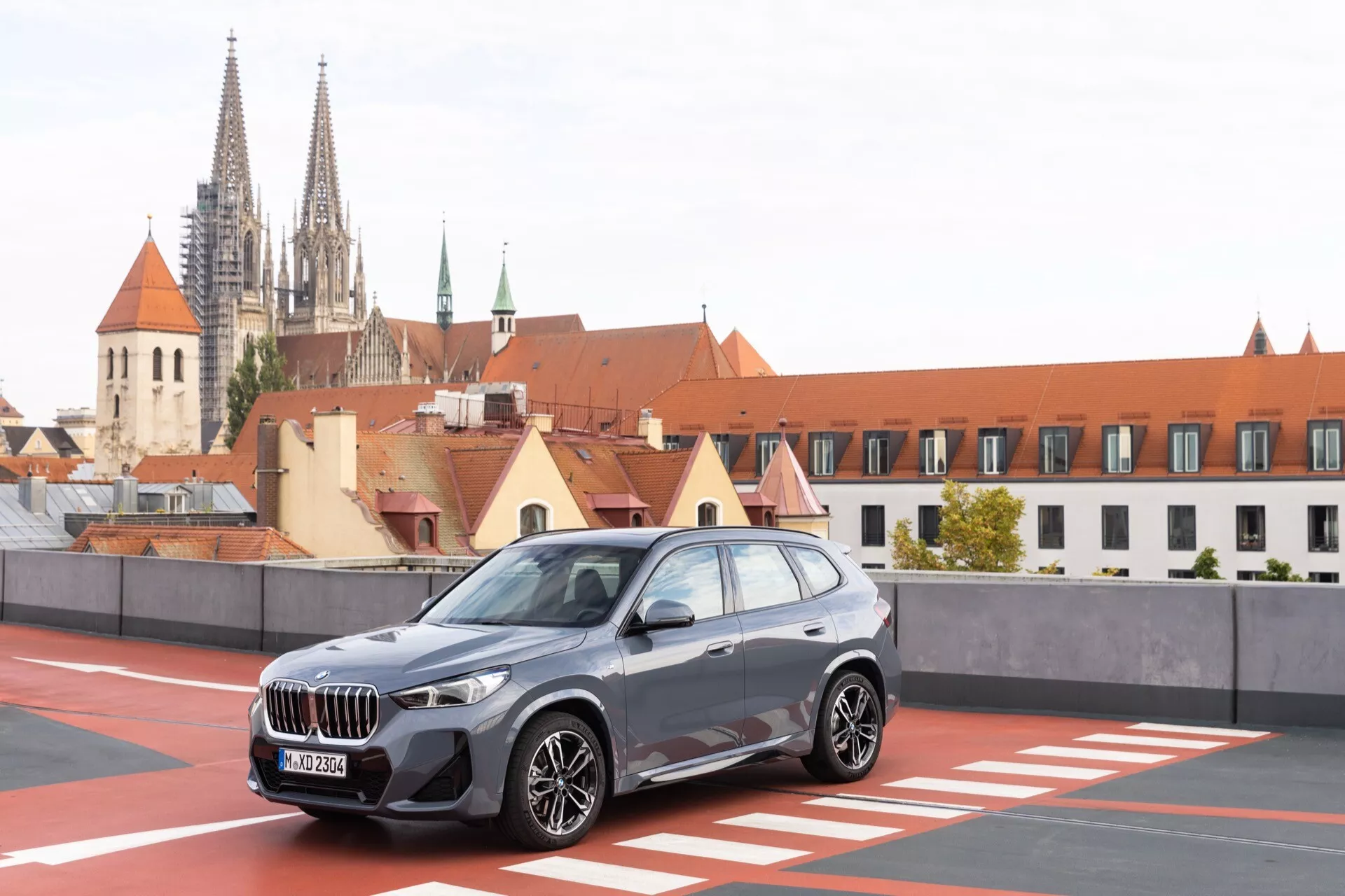 Bilden visar en BMW X1 2022 (U11) SUV parkerad, fotograferad från en trekvarts främre vy, som avslöjar dess framträdande frontgrill, vinkel-Led-strålkastare och aluminiumfälgar. Fordonets sidoprofil visar skulpterade linjer och synliga sidospeglar, vilket indikerar en lyxig design med potentiellt integrerade blinkers. Miljön verkar vara ett takparkeringsområde med en urban bakgrund innehållande historisk arkitektur, vilket kontrasterar modern fordonsingenjörskonst med ett klassiskt stadstema.