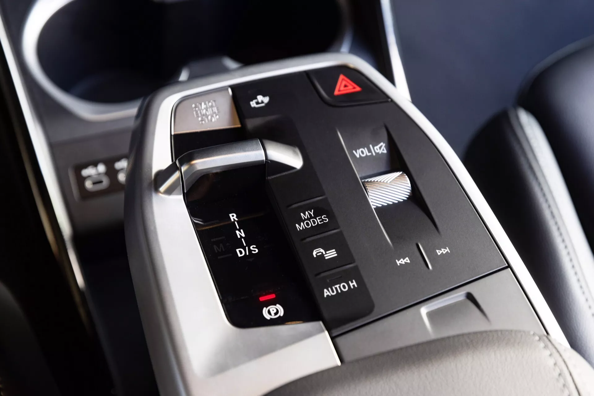 Närbild av mittkonsolen i BMW X1 2022 (U11) med en elektronisk växelväljare med etiketter 'R', 'N', 'D', 'P', media- och infotainmentskontroller, anpassningsbara 'MY MODES' och 'AUTO HOLD' funktioner, autostart/stoppfunktion och varningsljuskontakt, framhävande dess moderna, minimalistiska design och premiummaterial.