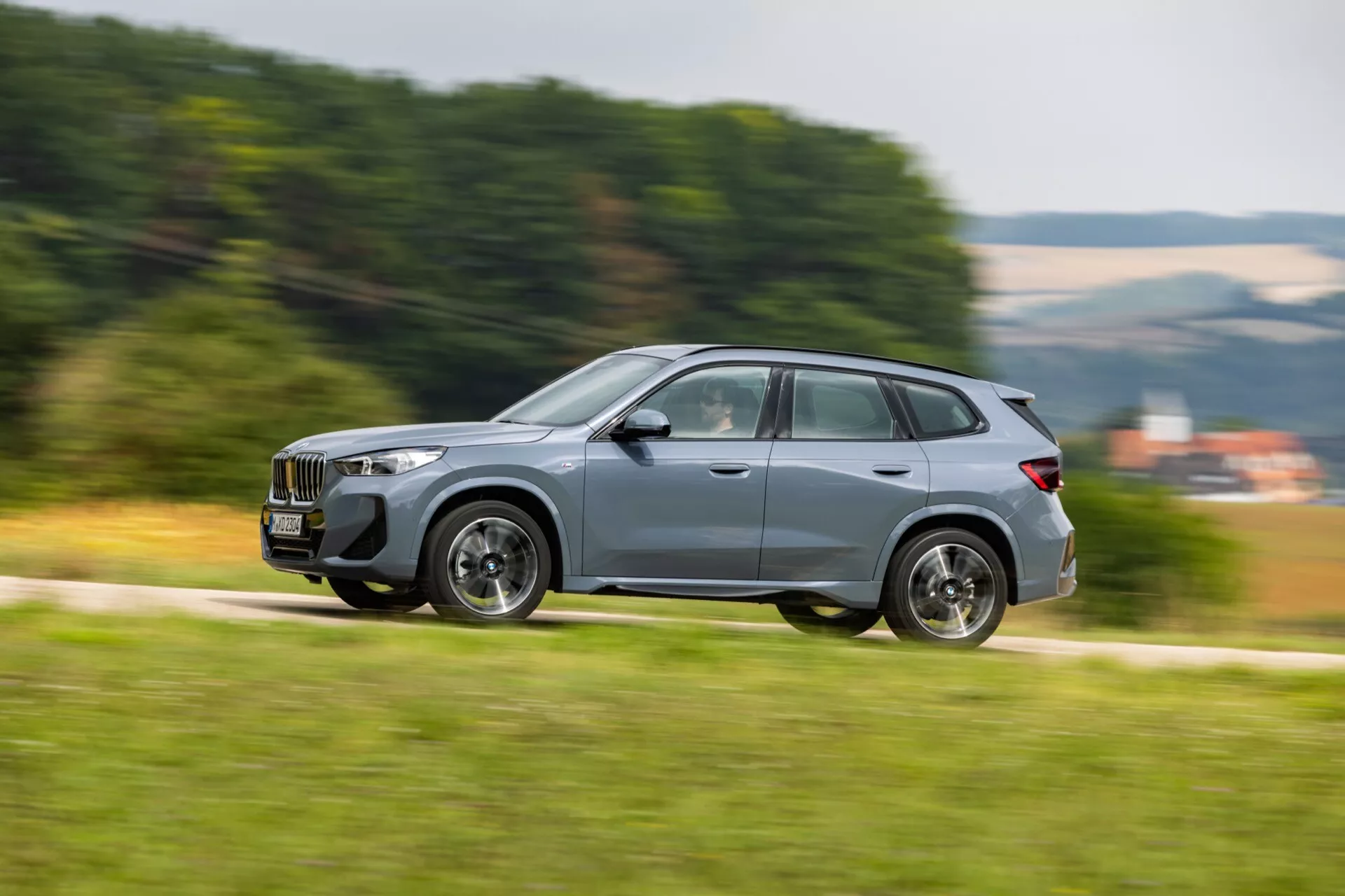 Dynamisk sidoprofilbild av en BMW X1 2022 (U11) i rörelse, som visar SUV:ens distinkta frontgrill, avancerad LED-strålkastardesign, hela sidovyn med karosserilinjer, rörliga hjul och kvalitetsfönsterreflektion, som demonstrerar markfrigång och rörelseoskärpa för en känsla av hastighet.