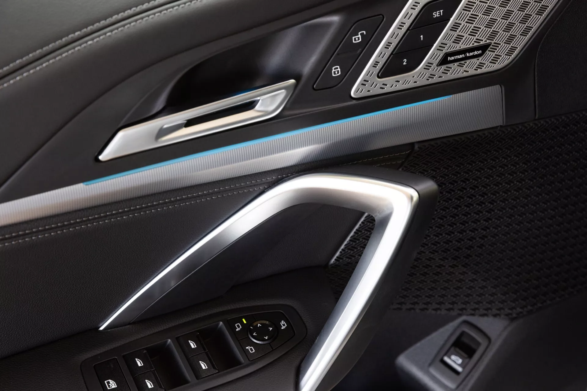 Närbild av förardörren på BMW X1 (U11) 2022 som framhäver ergonomisk design och teknikfunktioner inklusive kontroller för elektriska fönster, elektronisk justering av ytterspeglar, borstad metalldörrhandtag, stämningsbelysning och galler för högupplöst högtalare.