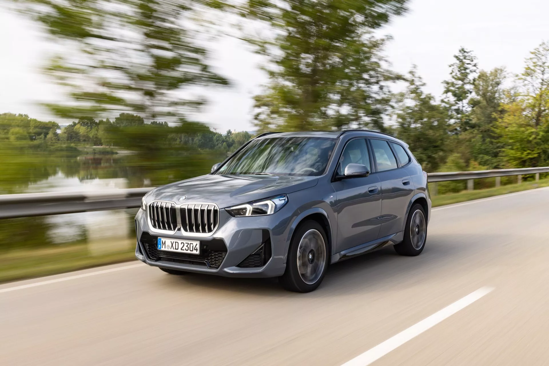 BMW X1 2022 (U11) i rörelse, fångad från en trekvarts frontal vinkel som visar fram- och sidoprofilerna. Synliga är den stora grillen med vertikala lameller, eleganta LED-strålkastare, stora luftintag i frontstöten, snurrande aluminiumfälgar med flerspokedesign och sidospeglar med integrerade blinkers. Bilen är en mörk, glansig SUV eller crossover med hög markfrigång, skulpterade aerodynamiska linjer, tonade sidofönster och en övergripande stilfull uppenbarelse, vilket skapar en känsla av hastighet och dynamisk körning.