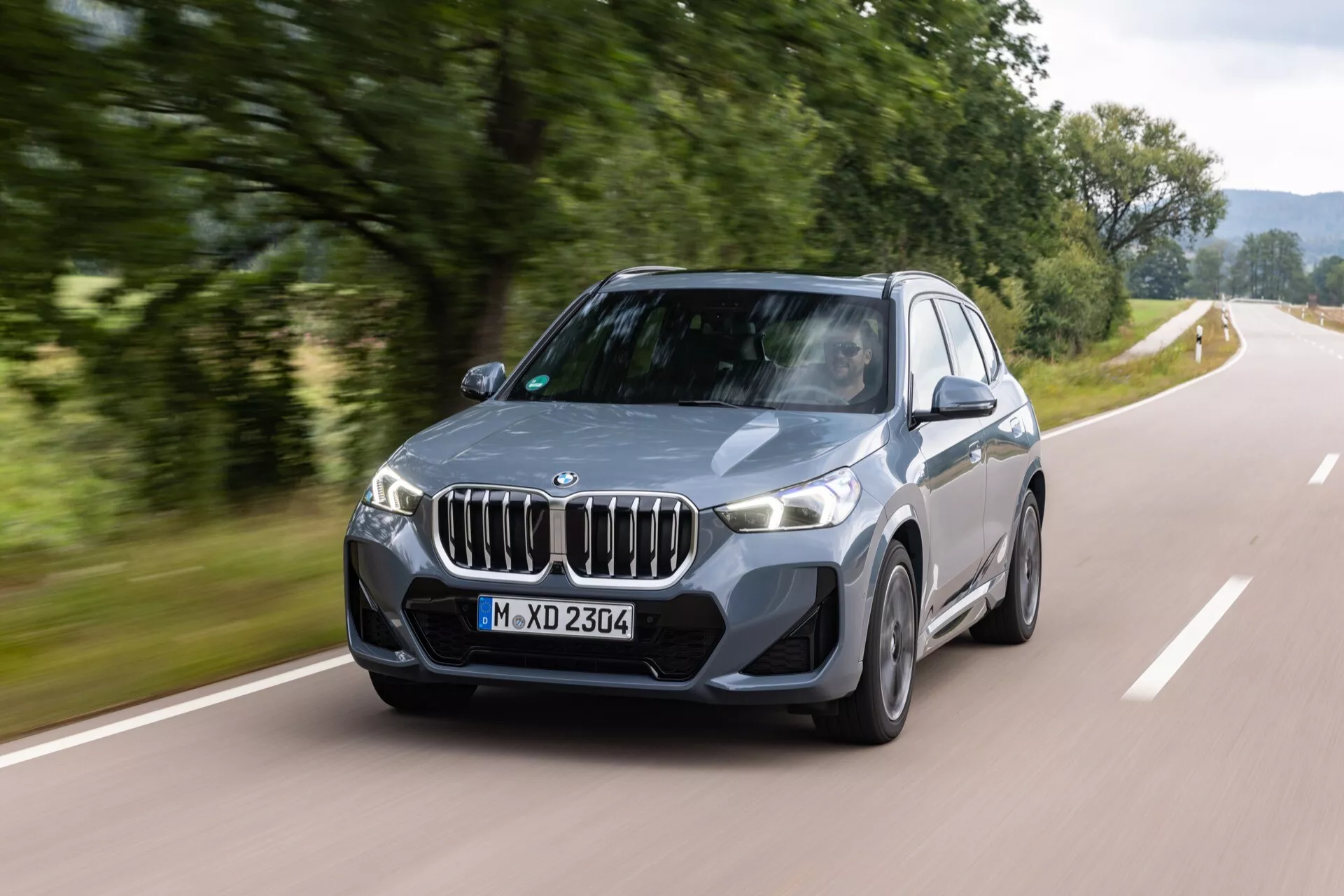 Grå BMW X1 (U11) från 2022 i rörelse på en landsväg med suddig grön landskapsbakgrund, visar sin främre tre fjärdedels vy, njurformad grill, LED-strålkastare, aggressiv stötfångarstil, flerspakiga legeringshjul, aerodynamiska linjer och sluttande taklinje som indikerar sportig design och avancerade teknikfunktioner.