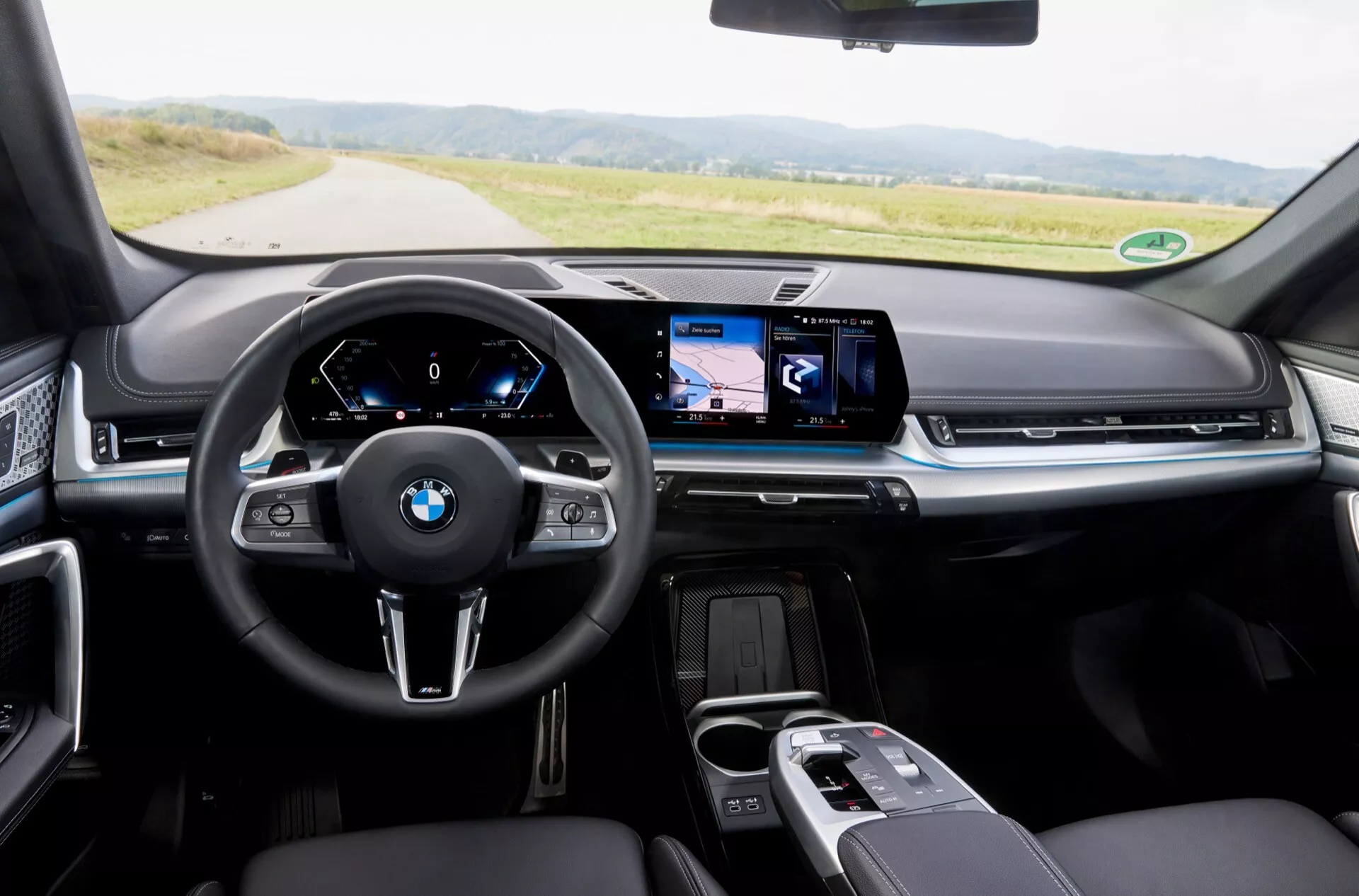 Interiörvy från förarsätet i BMW X1 (U11) 2022 som visar instrumentbrädan, BMW-emblemet på ratten och mittkonsolen med tekniska funktioner