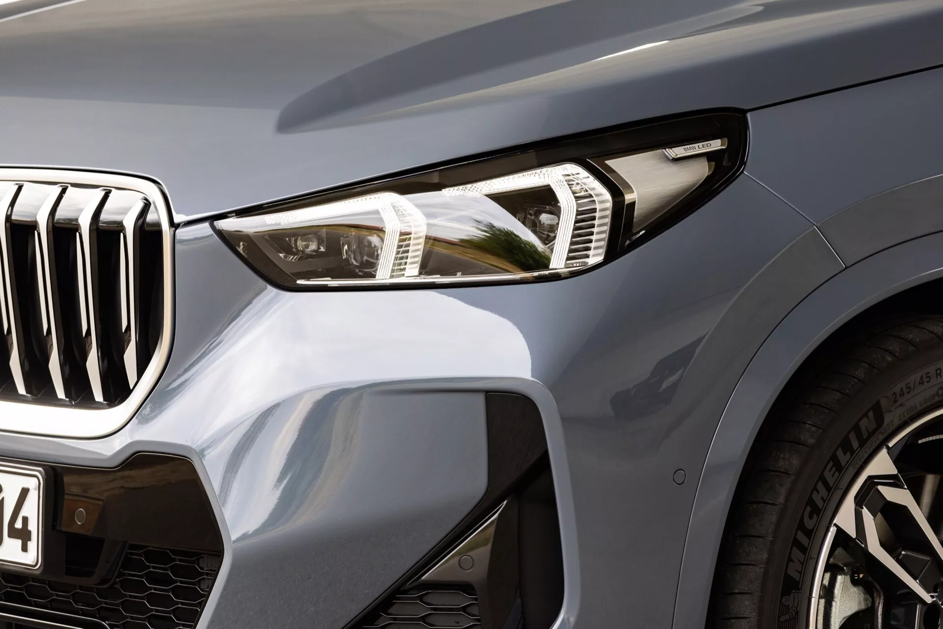 Närbild av vänster framsida av BMW X1 2022 (U11) som framhäver LED-strålkastaren med kurvljusfunktion, stor frontgrill med vertikala lameller, prestandaorienterade stora luftintag på stötfångaren, elegant metallisk lack och lågprofildäck på flerspakiga legeringshjul, som visar dess lyxiga och aerodynamiska design.
