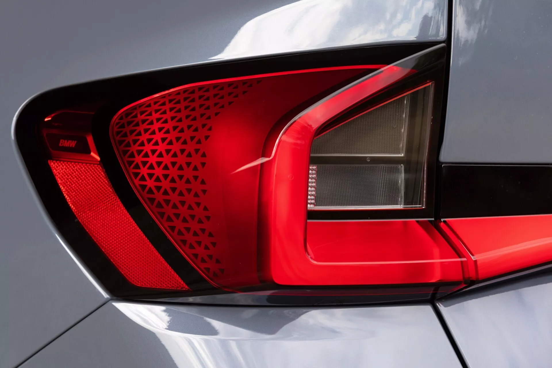 Närbild av bakljusmonteringen på BMW X1 2022 (U11). Bilden framhäver det moderna, övervägande röda bakljuset med ett geometriskt mönster för bättre ljusfördelning. Ett tydligt avsnitt för back- eller dimljus, LED-tekniken säkerställer effektivitet och ljusstyrka. Det blanka höljet sveper runt bilens sida och förbättrar synligheten från sidan. En subtil fenliknande utstickare längs den övre kanten antyder en aerodynamisk design. Idealisk för diskussioner om fordonssteknologi, design och säkerhetsfunktioner för fordon.