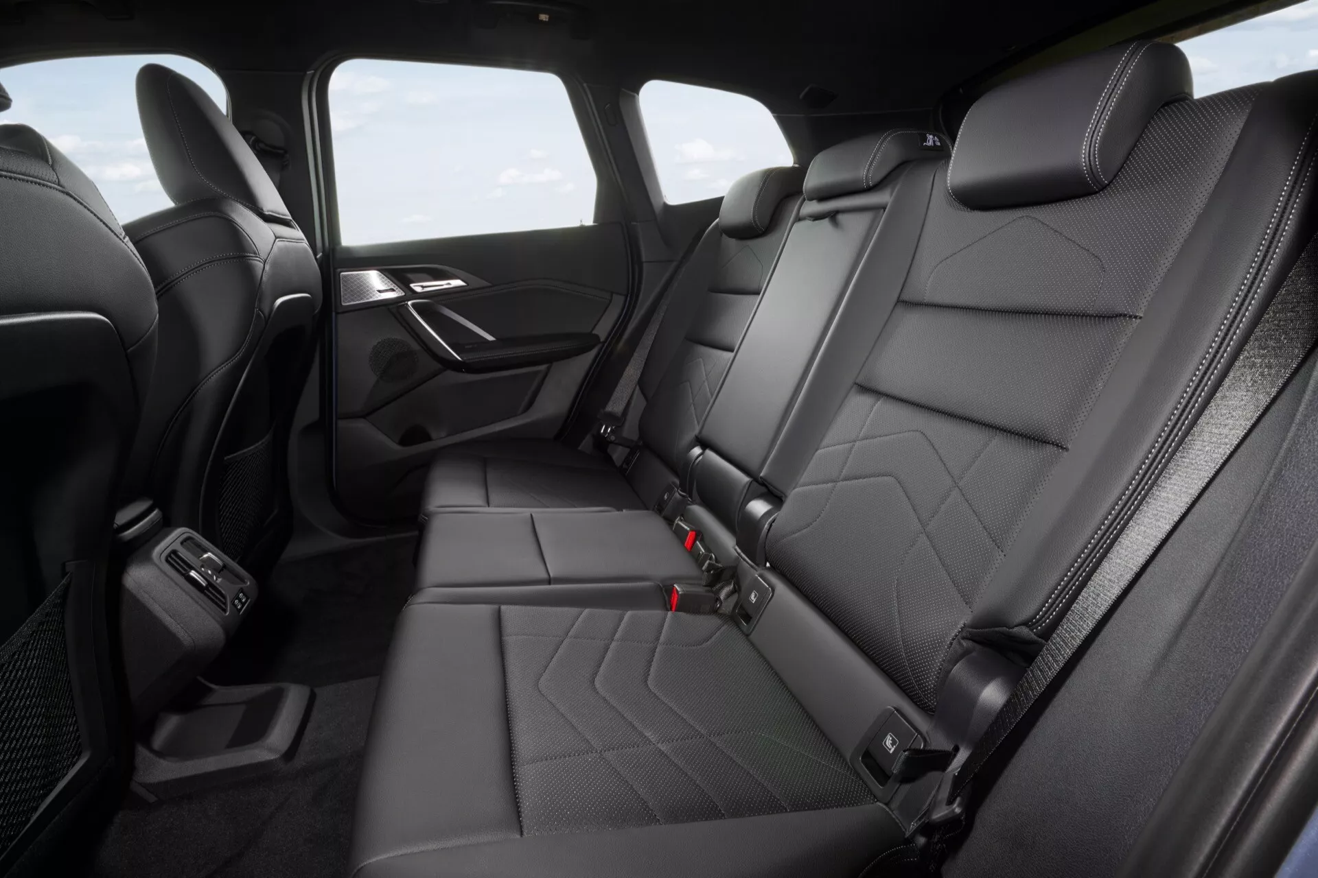 Baksätesområde i BMW X1 (U11) 2022 med quiltad läderklädsel, individuella nackstöd, synliga säkerhetsbälten, nedfällbart mittarmstöd, Isofix-punkter, dörrpanel med kromdetaljer, USB-port, luftkonditioneringens ventiler, mattbelagda golvet och justeringshandtag för säten, från en upphöjd vinkel