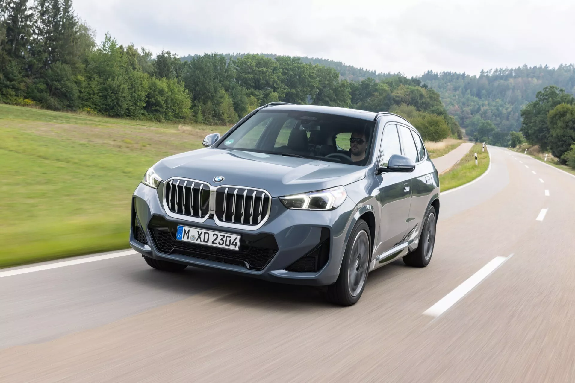 Lyxig SUV BMW X1 (U11) 2022 i rörelse med en grön, skogbeklädd landsbygdsbakgrund, som visar dess distinkta njurformade grill, eleganta LED-strålkastare, aerodynamisk frontstötfångare och flerspakiga legeringshjul från en främre trekvarts vinkel som betonar förarsidan.