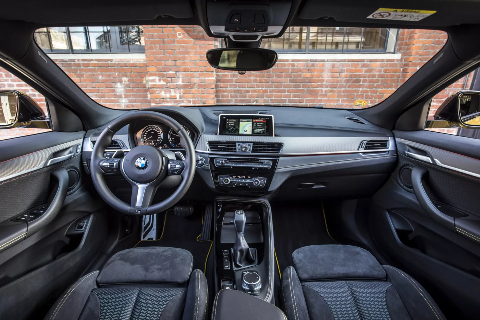Interiörvy av en BMW X2 2018, visar framkabinen från baksätets perspektiv med framhävning av ratten med monterade kontroller och BMW-logotyp, delvis dolt instrumentkluster, mittkonsol med växelspak och iDrive-system, icke-pekskärm för infotainmentskärm, lyxig instrumentpanel med trä eller metalldekor, dubbelzons klimatkontroll, konturerade framsäten, praktiska mugginnehållare och förvaringsutrymmen samt dörrpaneler med kontrollfunktioner under ett solbelyst takfönster.