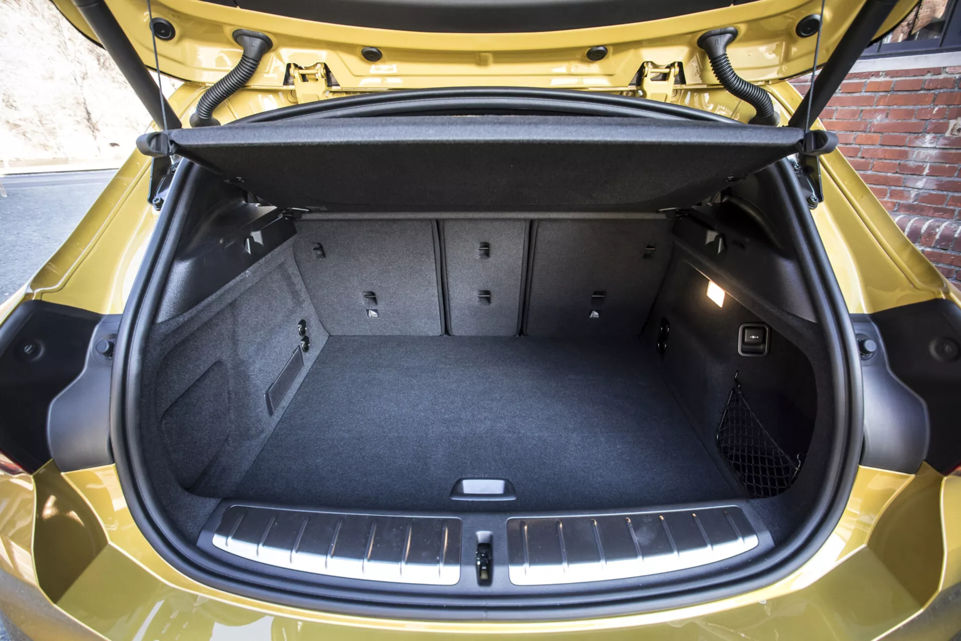 Närbild av öppet bagageutrymme på BMW X2 2018 som framhäver ett tomt, rent lastutrymme med nätad ficka för säkerhet av föremål, platt lastgolv och skyddande lining, lock för förvaring under golvet, reflekterande remsa under dörren, i en glansig gul finish