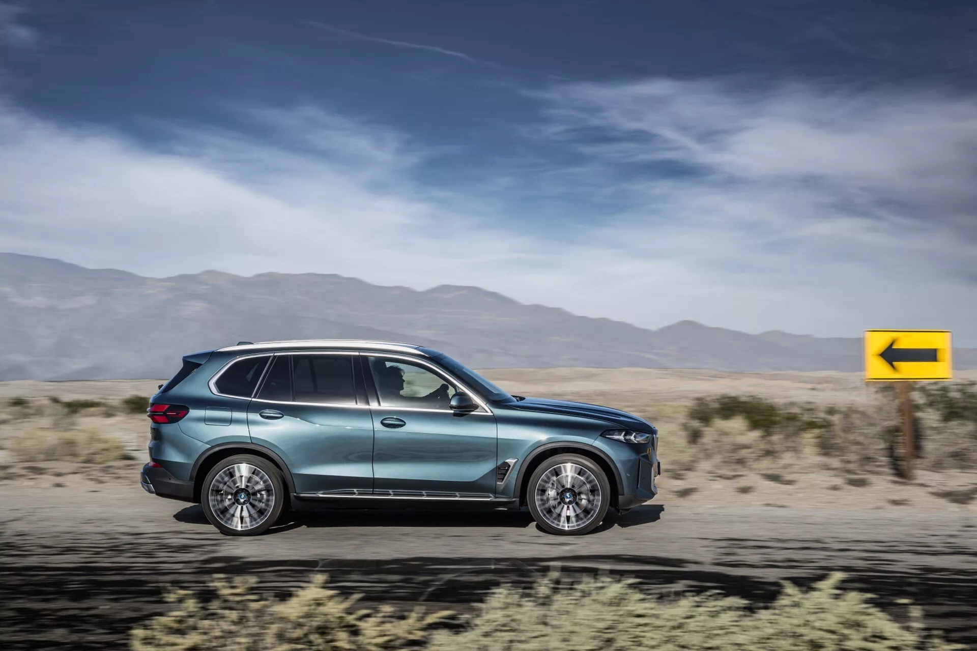 SUV BMW X5 2023 (G05-F95-Facelift) i rörelse, visar aerodynamik, flerspåriga legeringshjul, integrerade dörrhandtag, aerodynamiska sidospeglar, tonade fönster, sidoblinkers, metallisk lack och modern baklyktedesign mot en suddig ökenbakgrund.