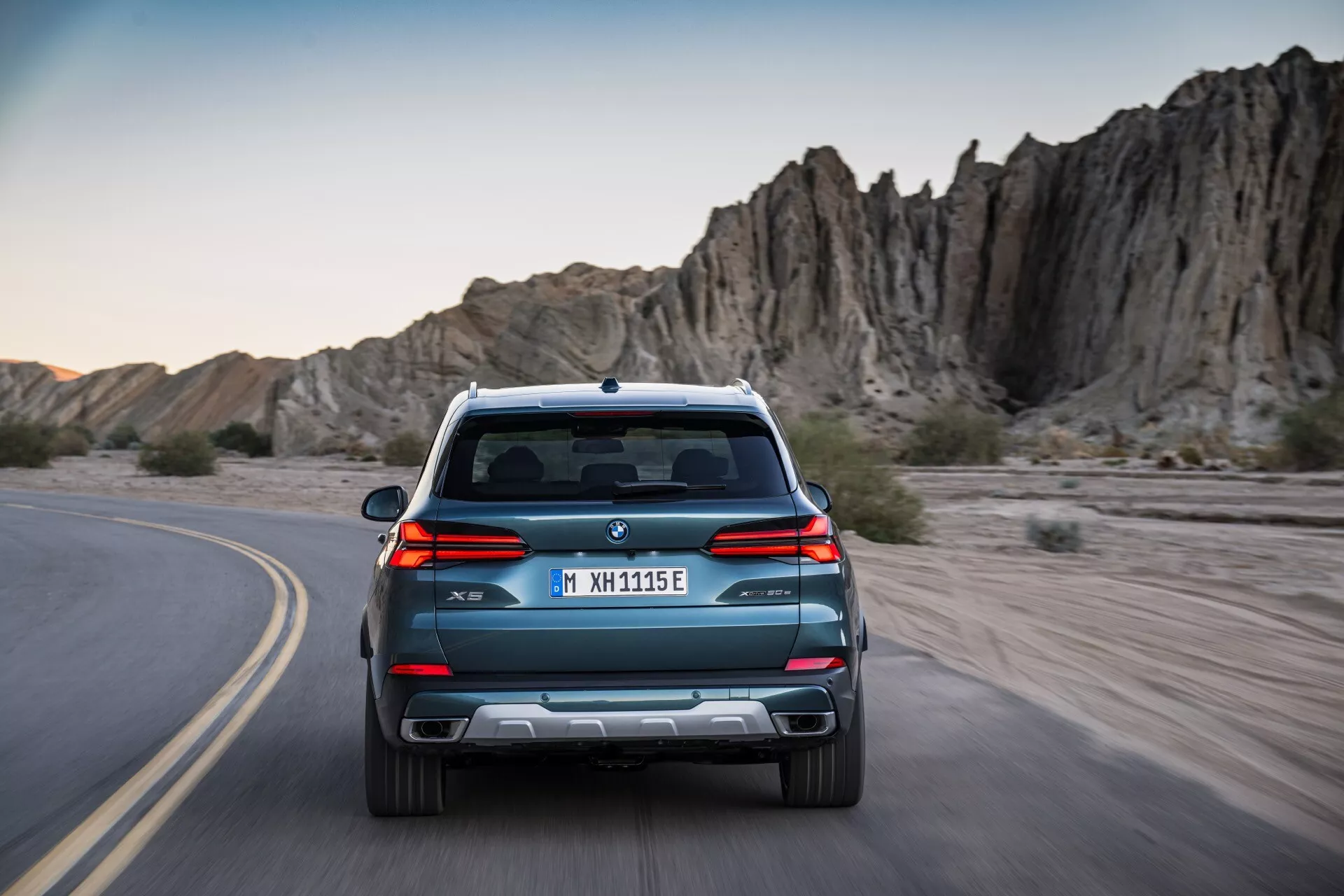 Bakvy av BMW X5 G05 F95 facelift 2023, kör på en kurvig väg, med snygga linjer, dubbla avgassystem och parkeringssensorer mot en äventyrlig klippig terräng i bakgrunden