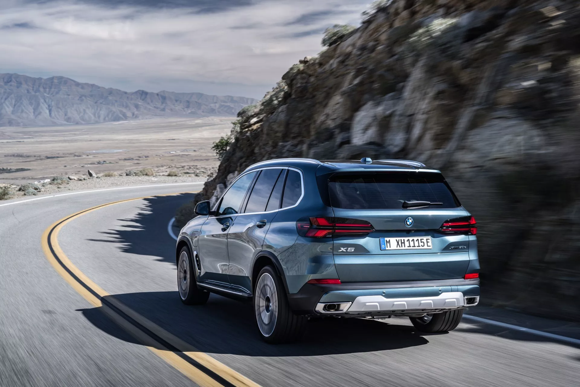 Dynamiskt bakre trekvartsutsikt av BMW X5 2023 (G05-F95 Facelift) på en kurvig bergsväg som visar dess snygga baklucka, sportiga spoiler och lyxiga legeringshjul