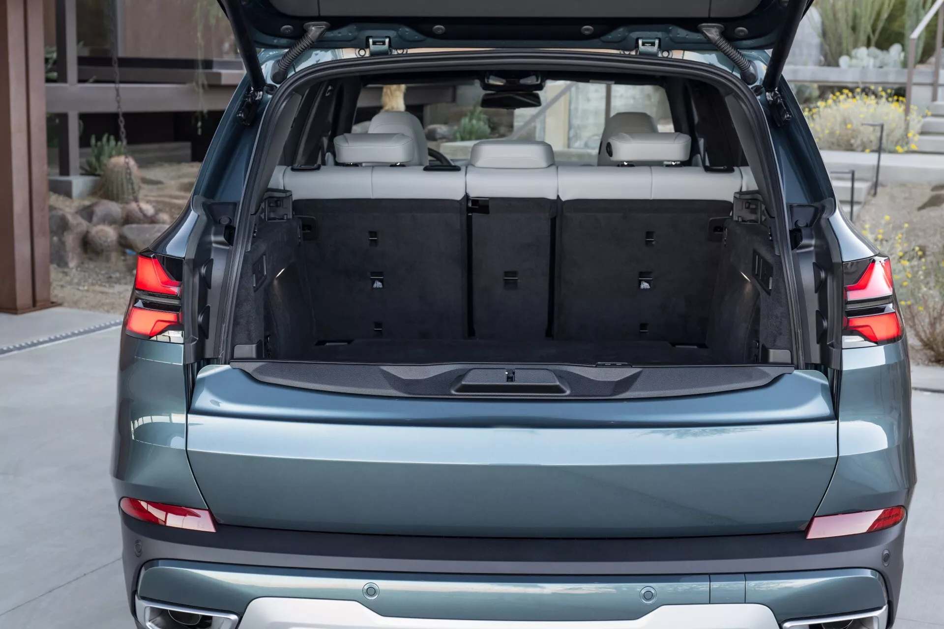Bakvy av BMW X5 (G05-F95 Facelift) 2023 som framhäver det rymliga öppna bagageutrymmet, moderna horisontella bakljus och bakre designfunktioner inklusive parkeringssensorer och kromlist, med en glimt av den tre sätes konfigurationen i andra raden.