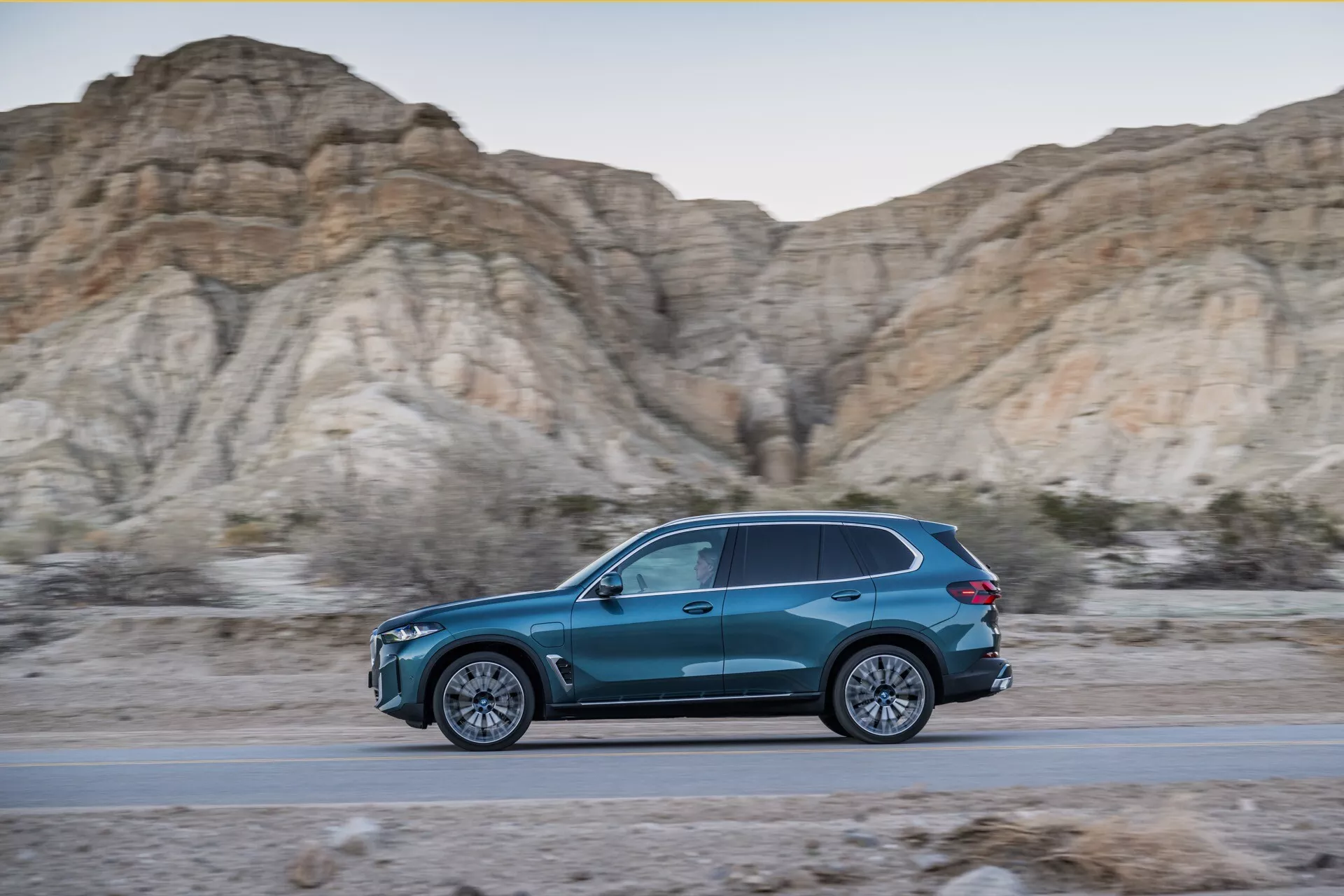 Dynamisk sidoprofil av BMW X5 2023 (G05-F95 ansiktslyft) i rörelse, blå SUV i tårform på en väg med bakgrund av karga, kala kullar under gyllene timmen, visar en slank design, aerodynamisk effektivitet, flerspakiga legeringshjul och LED-belysning