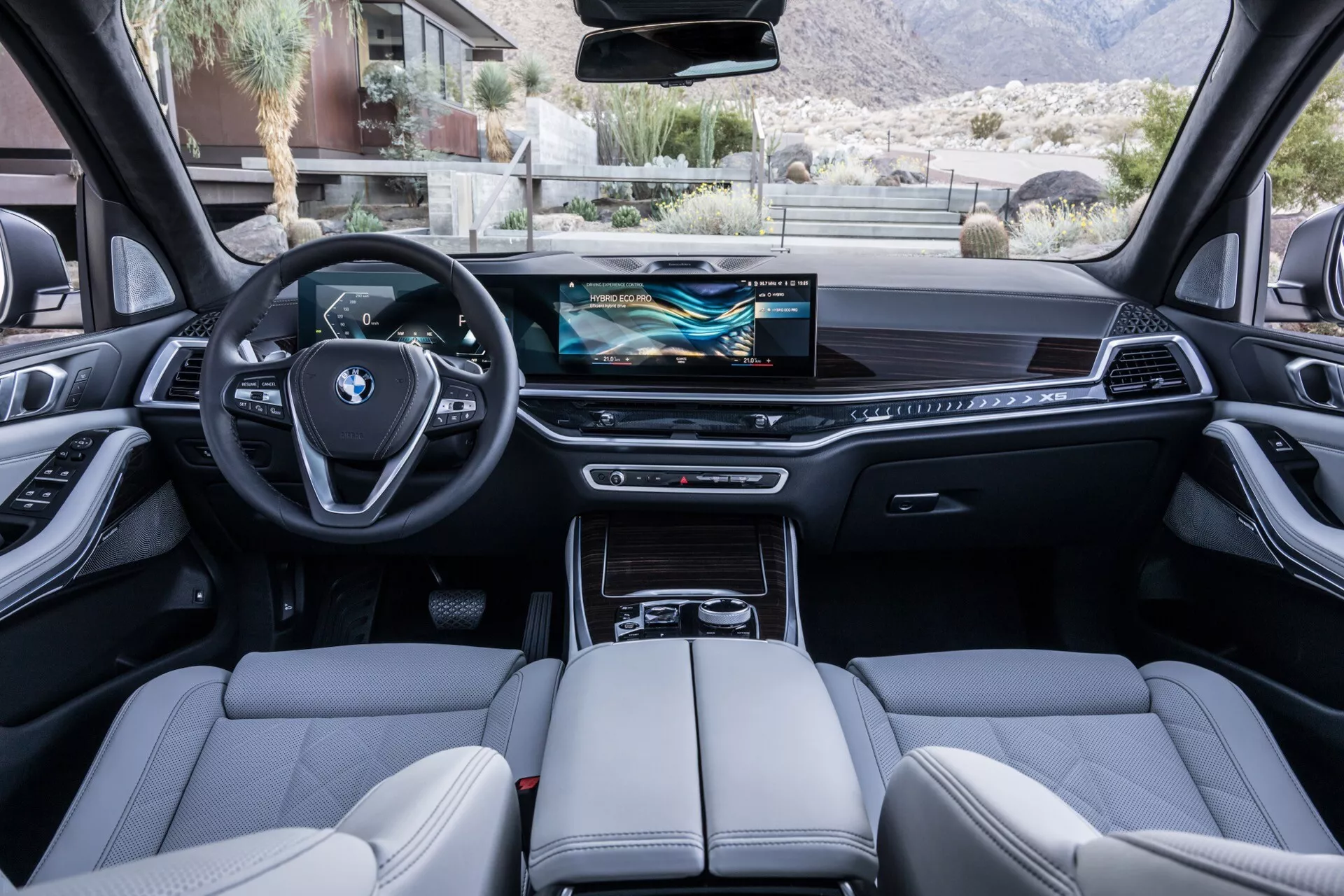 Interiörvy av BMW X5 2023 (G05-F95 Facelift) från baksätet som visar en sportig trespoke ratt med integrerade multifunktionsknappar, digital instrumentpanel, bred infotainment pekskärm, minimalistiska kontroller på instrumentbrädan, elektronisk växelväljare, högkvalitativa lädersäten med detaljerade sömmar, panoramasoltak och avancerade teknologiska funktioner