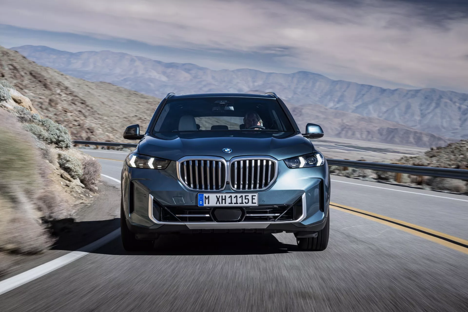 BMW X5 2023 (G05 F95 ansiktslyftning) i rörelse, fångad från en framifrån tre kvarts vinkel som belyser dess LED-strålkastare, njurgrill med aktiva luckor, legeringshjul och det smidiga aerodynamiska designen mot en bakgrund av bergsöken, vilket antyder avancerad teknik och potentiell elektrisk eller hybrid kapacitet