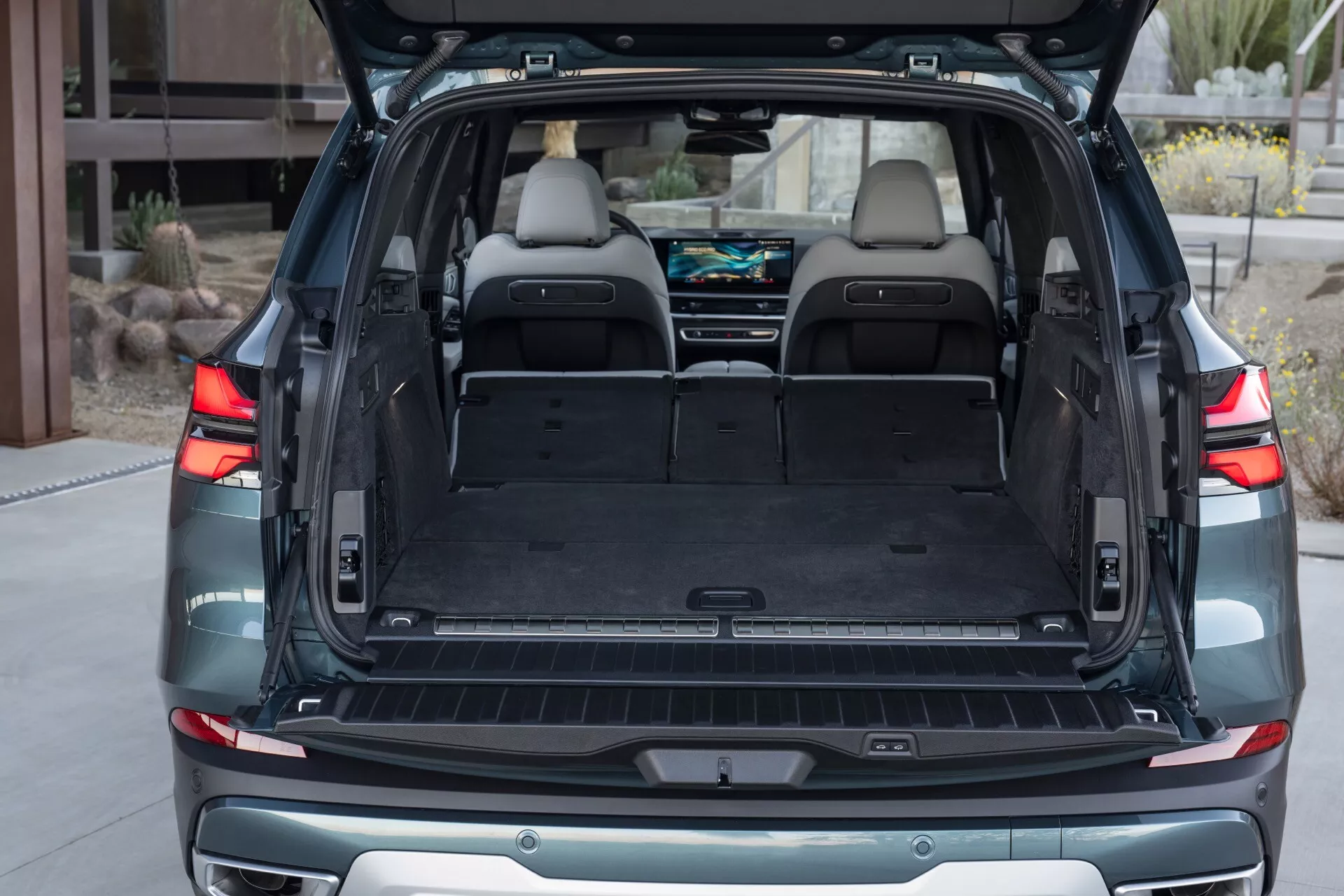 BMW X5 2023 (G05-F95 Facelift) visar ett rymligt och välbelyst bagageutrymme med plant lastgolv, upprätt andra radens säten, lyxig inredning i ljusläder, synliga lastfästpunkter och 12V-uttag, tillsammans med ett stötskydd på stötfångaren i en bostadsmiljö