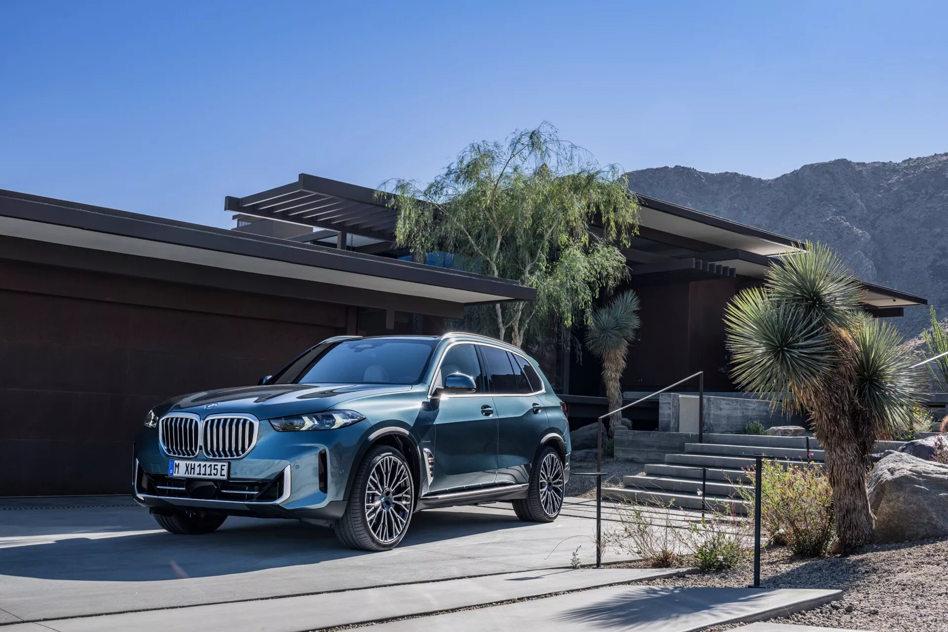 Lyxig SUV BMW X5 2023 (G05-F95 Facelift) parkerad framför ett modernt hus med öken och bergslandskap i bakgrunden, med metallisk finish, stor grill, LED-strålkastare och flerspakiga aluminiumfälgar