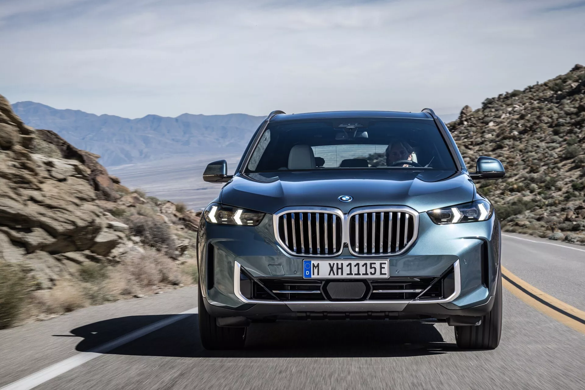 Modernt lyxigt SUV BMW X5 2023 (G05-F95-Facelift) i rörelse, med stor njurformad grill, slanka LED-strålkastare och avancerade teknikelement som framåtriktad kamera, avbildad från en främre trekvarts vinkel som visar fordonets dynamiska hållning och design detaljer såsom skulpterade huven och skärmarna.
