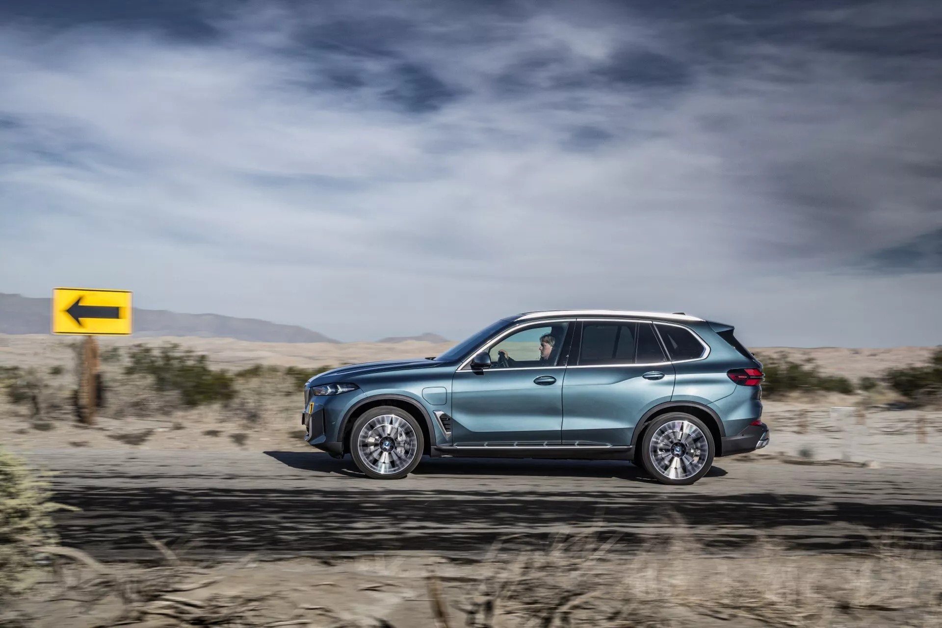 Sidoprofil i rörelse av BMW X5 2023 (G05-F95 Facelift), som visar dess metalliska lack, aerodynamisk design, lyxiga legeringshjul, tonade fönster med blinkers i speglarna, takräcken och avancerad LED-teknik, mot en ökenliknande bakgrund med en dynamisk gul vänstersvängskylt.