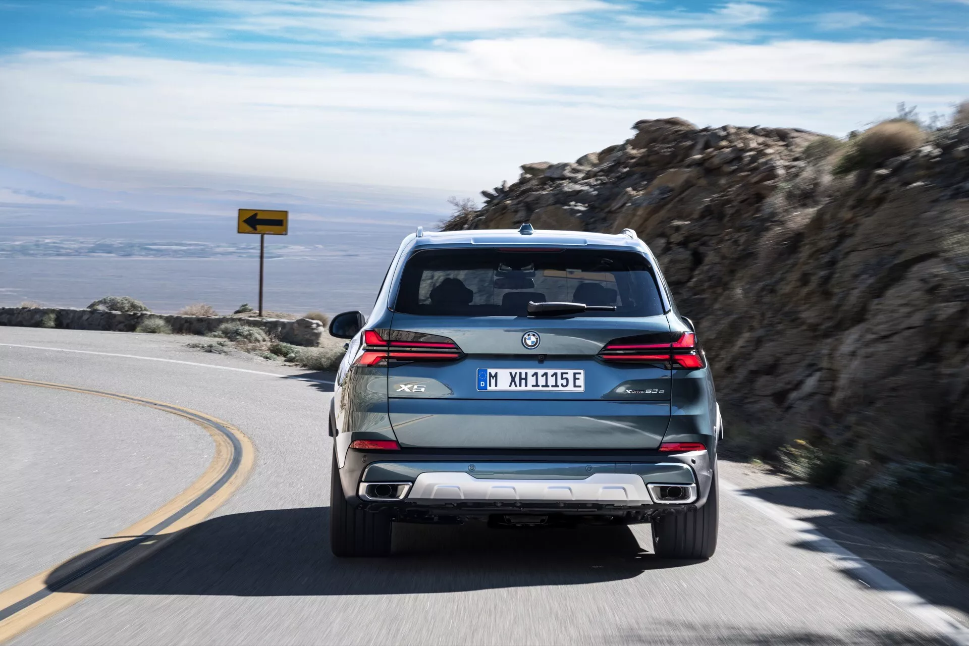 2023 BMW X5 (G05-F95) facelift xDrive45e med M Sport-paket som kör iväg på en bergsväg, visar upp sin PHEV-status med 'eDrive' och 'M'-märken, dubbla avgasrör, LED-baklyktor och europeisk registreringsskylt.