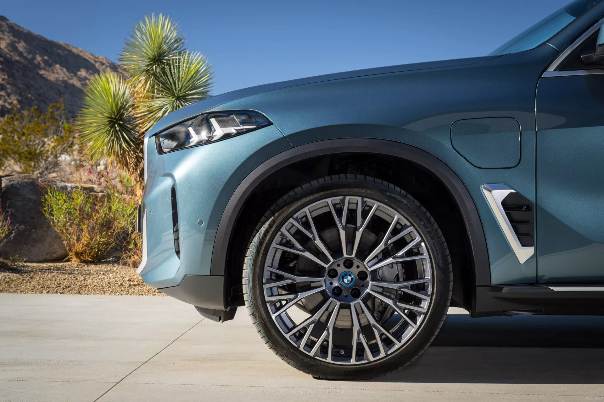 Närbild av BMW X5 (G05-F95) facelift 2023 som framhäver framhjulet och skärm med avancerade fjädringssystem, tvåtonigt stjärnformat lättmetallhjul och prestandainriktad lågprofildäck, visande sportig aerodynamisk design och parkeringsassistans teknologi mot en dagtid ökenbakgrund.