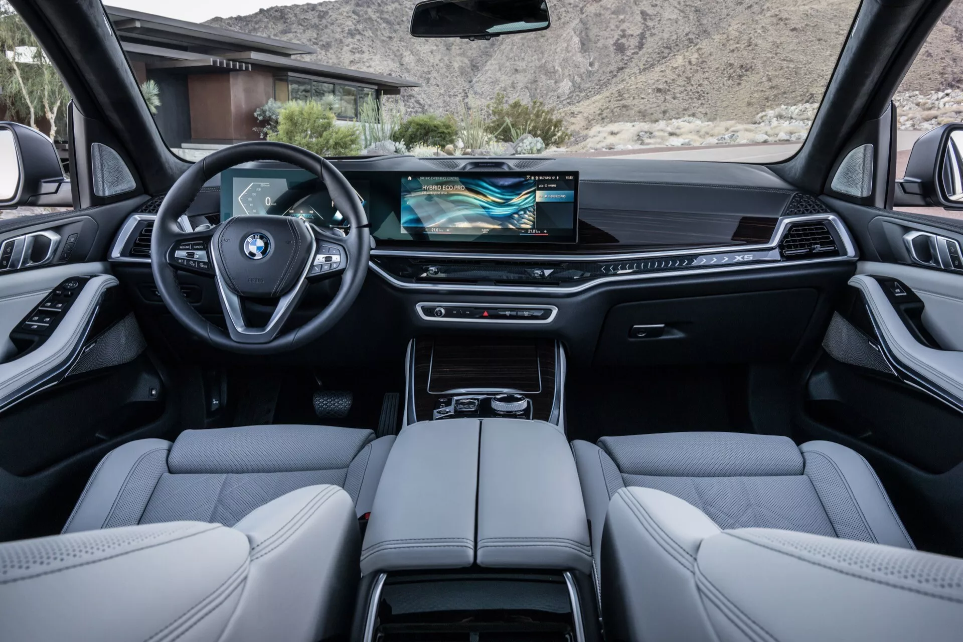 Interiörvy av BMW X5 2023 (G05 F95 ansiktslyft) från baksätet med fokus på instrumentbrädan med widescreen infotainment-display, läderinbunden multifunktions sportratt och lyxiga avslut inklusive fina lädersäten och metalliska accenter