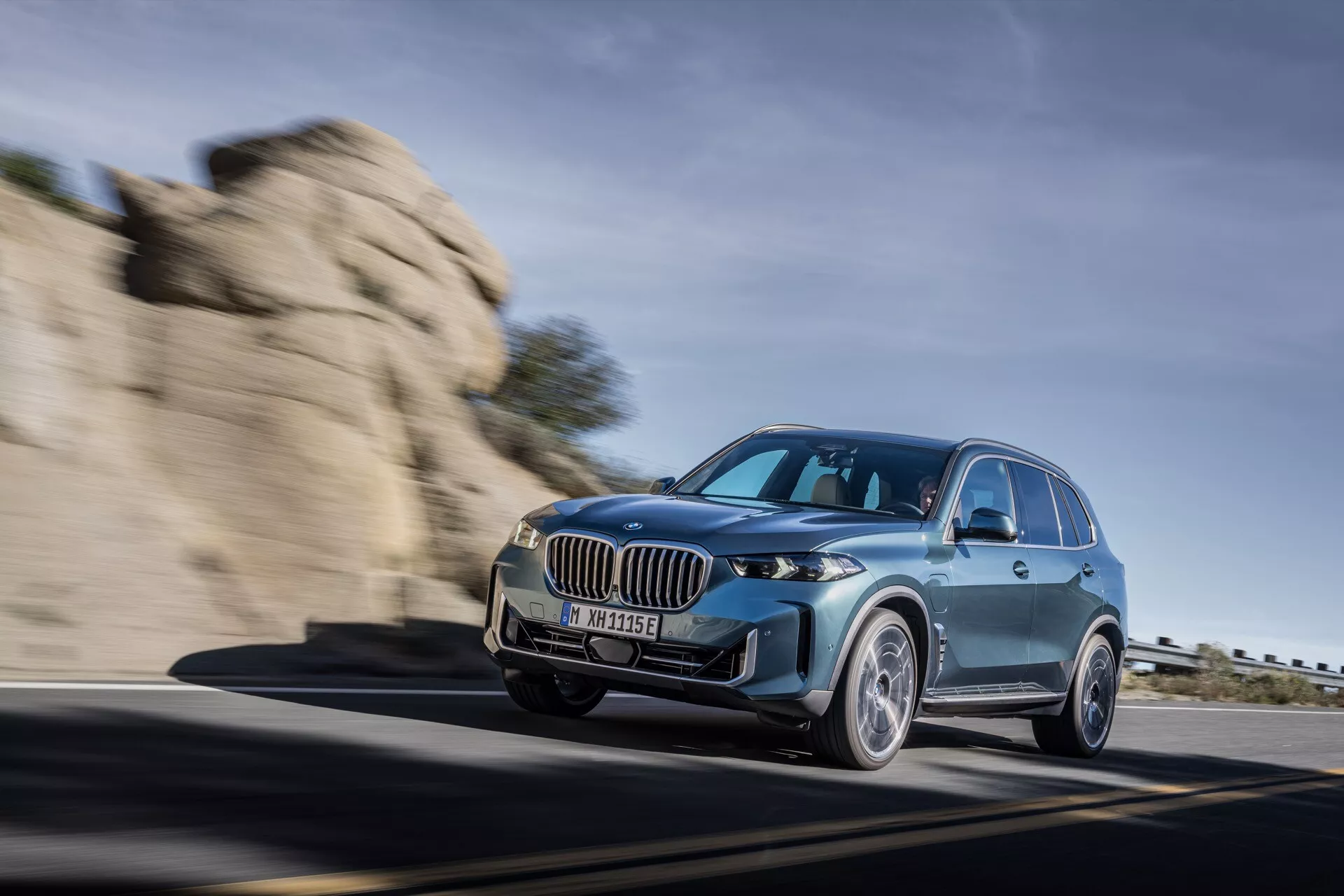 Lyxig SUV BMW X5 2023 (G05-F95 Facelift) i rörelse, visas upp i en dynamisk körscen på en bergig väg med en låg främre trekvarts vy som framhäver dess snygga LED-strålkastare, stora njurgrill och aerodynamisk design.