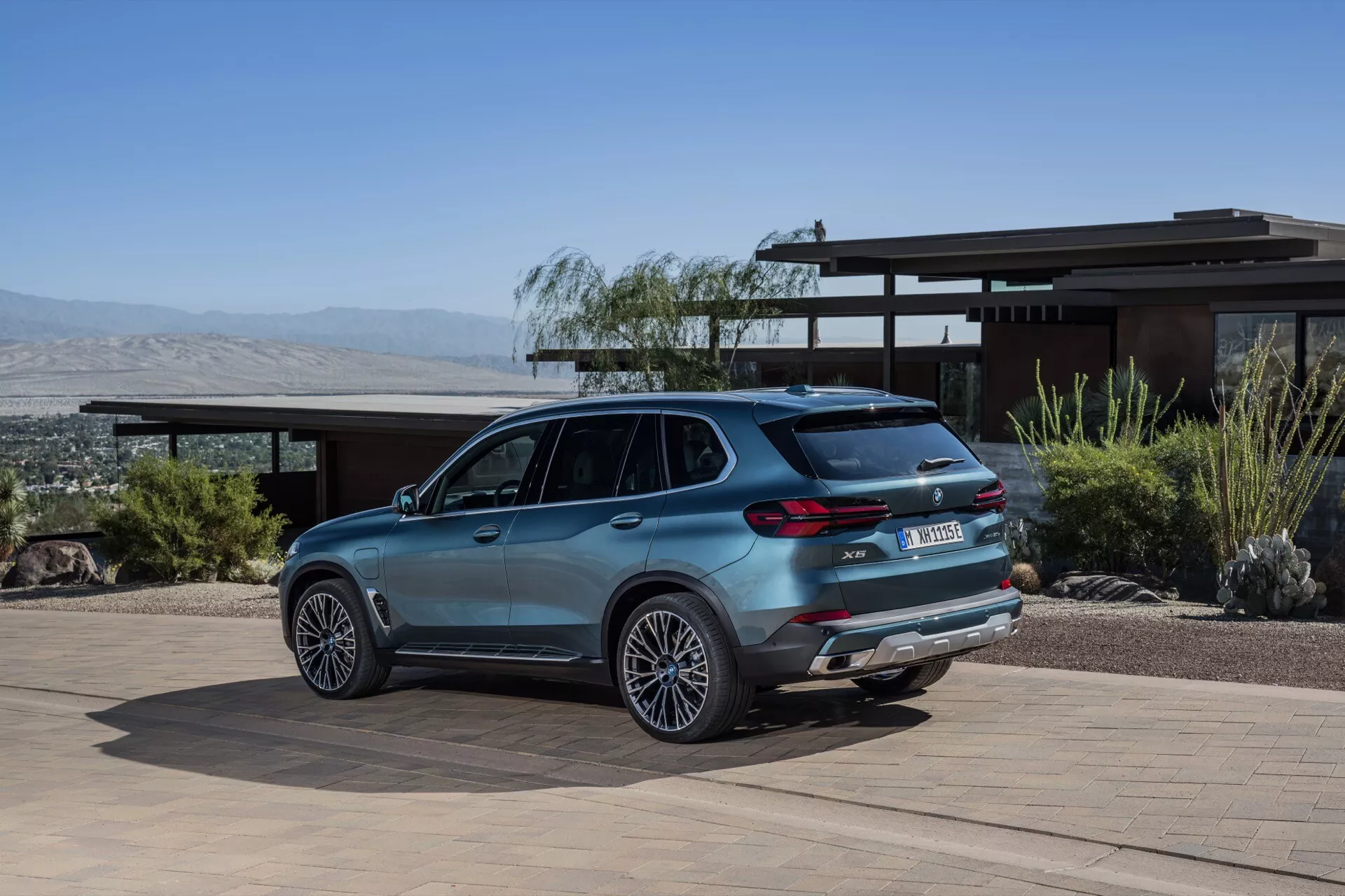 Lyxig SUV BMW X5 2023 (G05-F95 Facelift) parkerad på en bostadsuppfart med ökenlandskap och berg i bakgrunden, visar legeringshjul, dubbla avgasrör, LED bakljus, bakspoiler och takräcken från ett trekvarts bakvinkel.