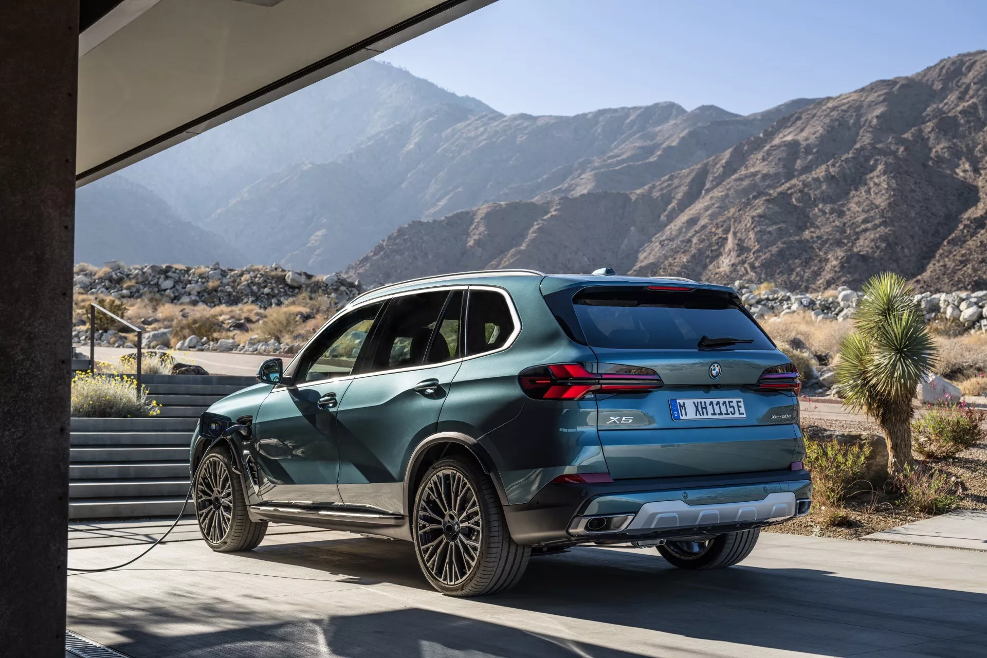 Profilvy av BMW X5 2023 (G05-F95-Facelift), som framhäver dess aerodynamiska design, moderna hjuldesign och laddningsfunktion, mot en modern struktur och bergslandskap, betonar dess miljövänliga elektriska eller hybrida natur och mångsidiga miljökapaciteter.