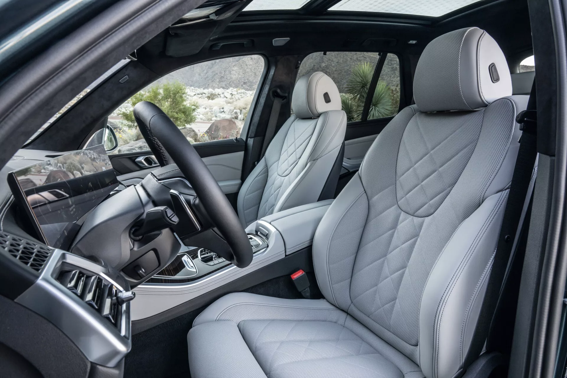 Interiörvy av BMW X5 2023 (G05 F95 ansiktslyft) genom öppen framdörr på passagerarsidan som framhäver en lyxig förarstol med ergonomisk quiltad klädsel, avancerad mittkonsol med växelspak och metalliska accenter, multifunktionell ratt, och del av instrumentbrädan med underhållningssystem
