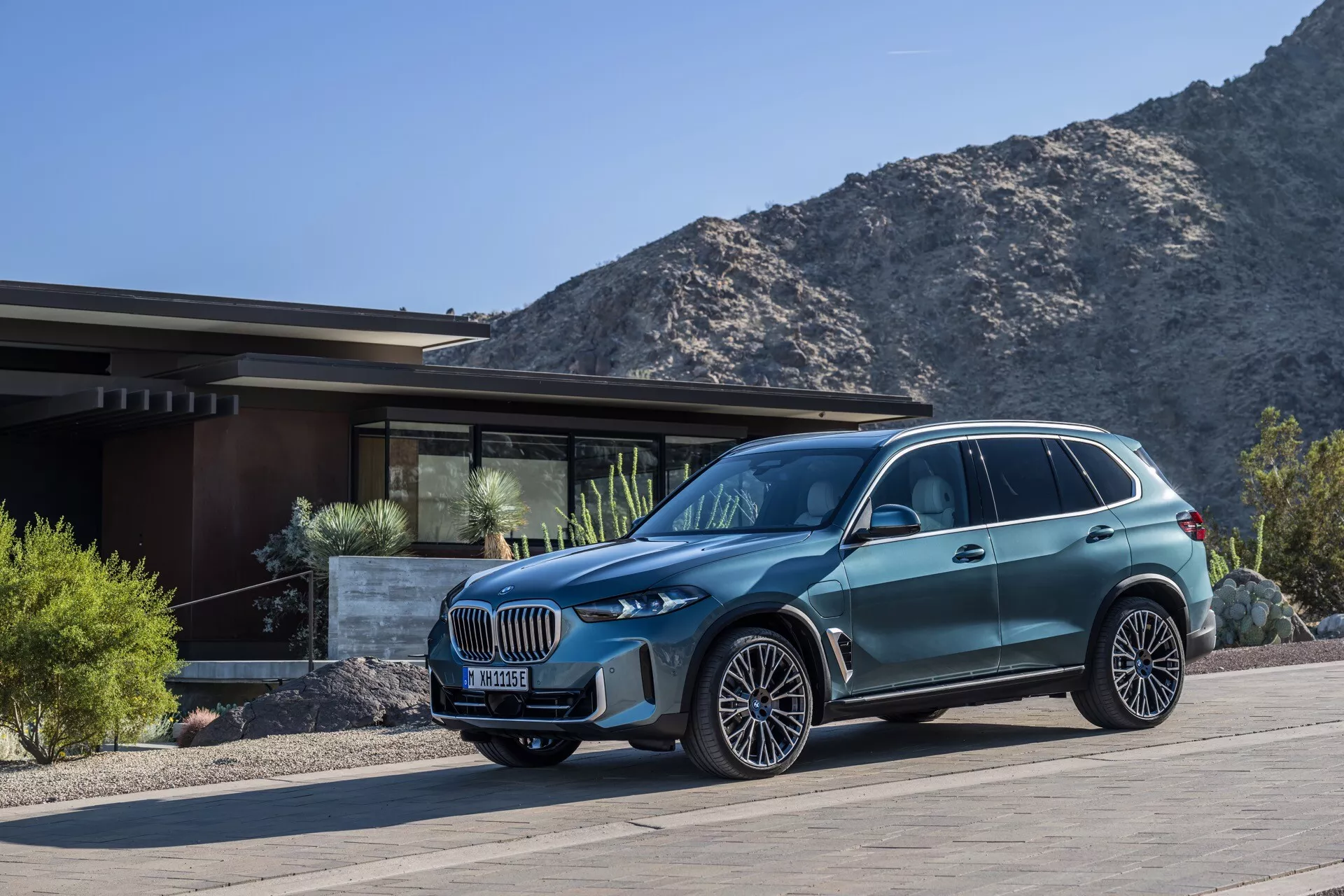 Trekvarts framsida av en lyxig crossover SUV BMW X5 2023 (G05-F95-Facelift) framför en exklusiv bostadsbyggnad med berg i bakgrunden, som framhäver dess stora njurformade grill, adaptiva LED-strålkastare, flerspakiga legeringshjul, dynamiska karosslinjer och avancerade designfunktioner.