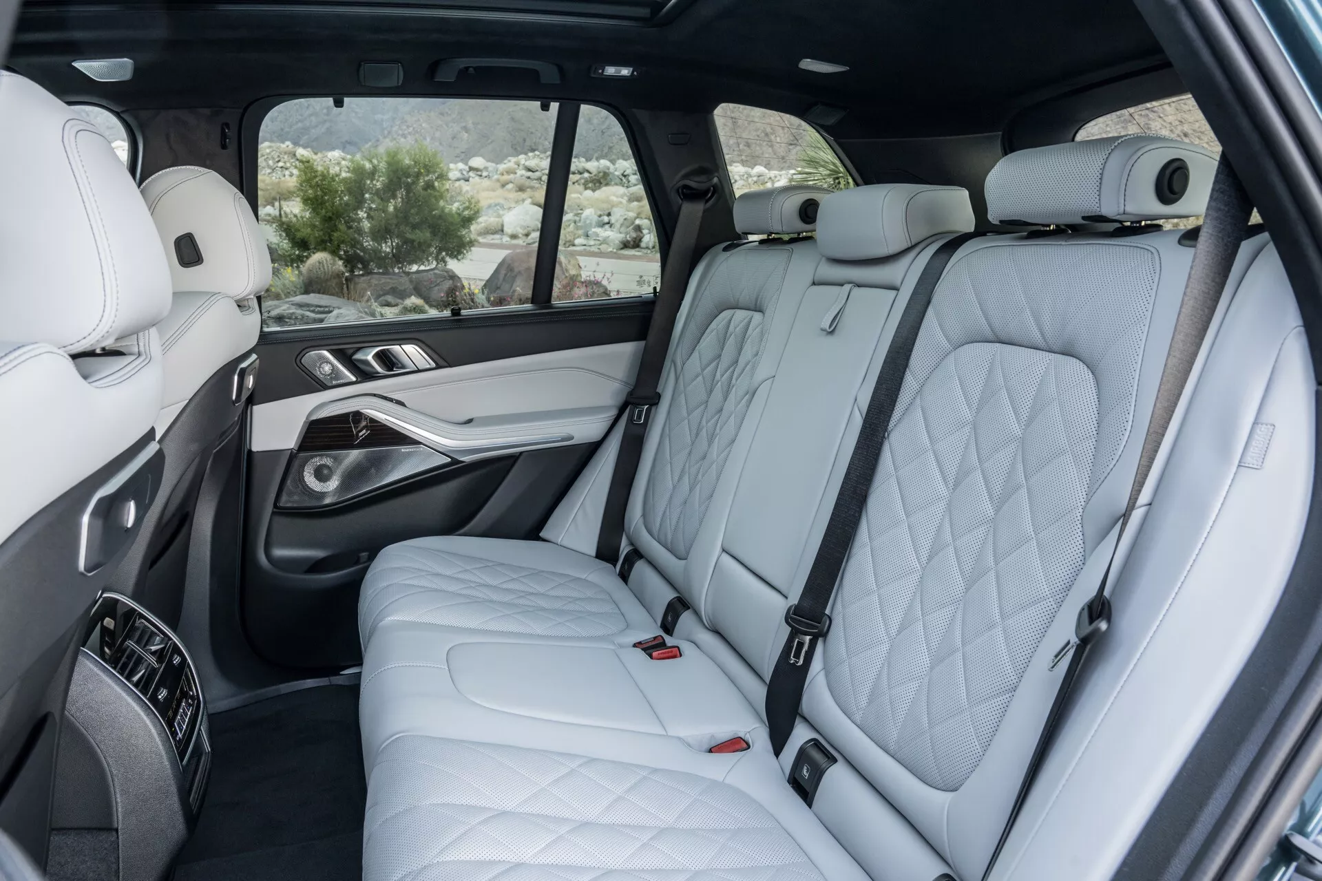 Interiörperspektiv av BMW X5 2023 (G05-F95-Facelift) som framhäver lyxiga andra radens säten med quiltat läder, premium dörrpanel med trä- och metallaccent, bekvämligheter i mittkonsolen, panoramasoltak, elektriska kontroller för bakre säte och avancerade synlighetsfunktioner