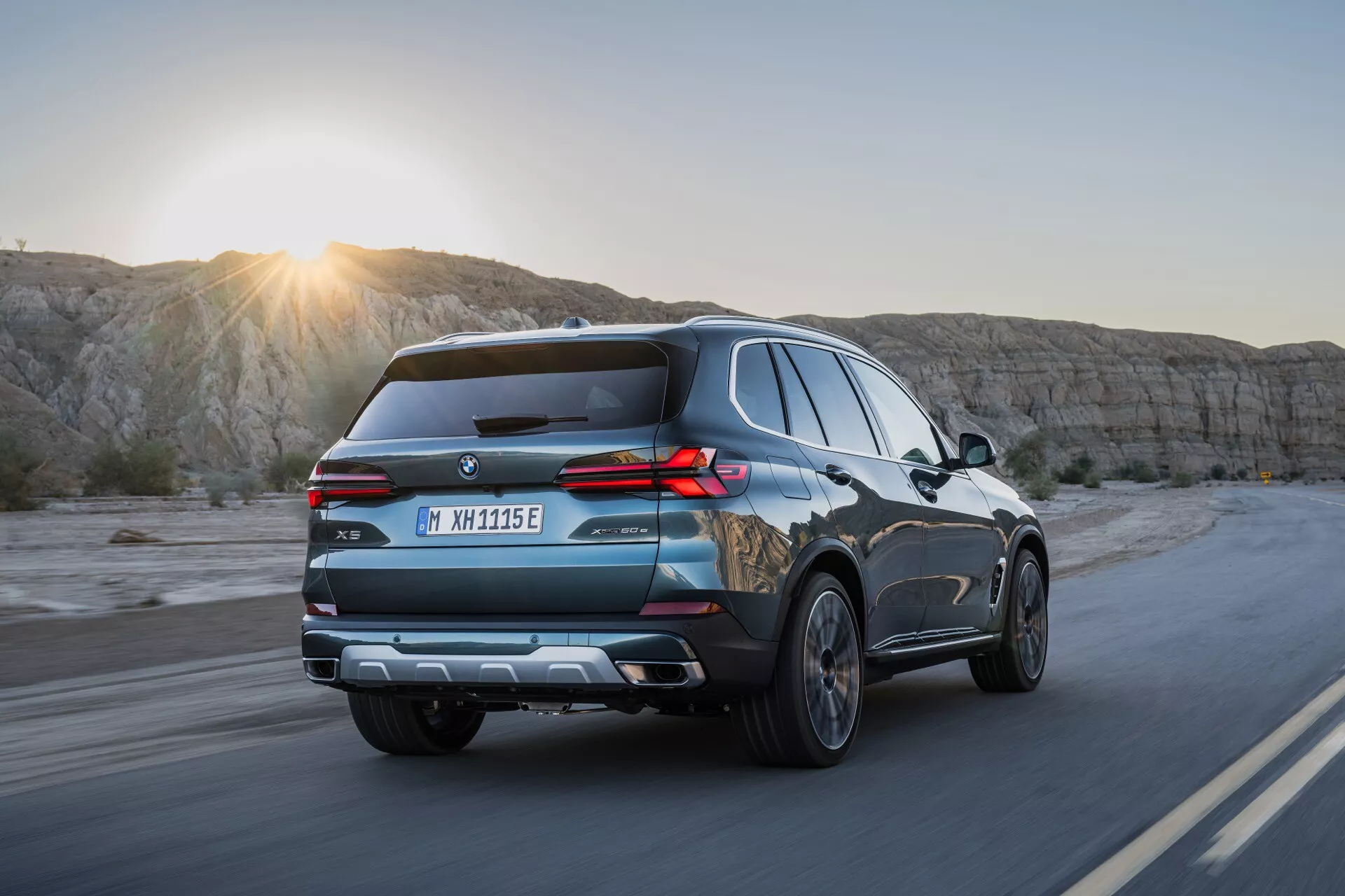 Modern SUV BMW X5 2023 (G05-F95-Facelift) i rörelse på en naturskön motorväg vid soluppgång eller solnedgång, fångar dynamisk baktre kvarts vinkel med LED-bakljus, takräcken och avancerade teknikfunktioner under mildt solglitter.