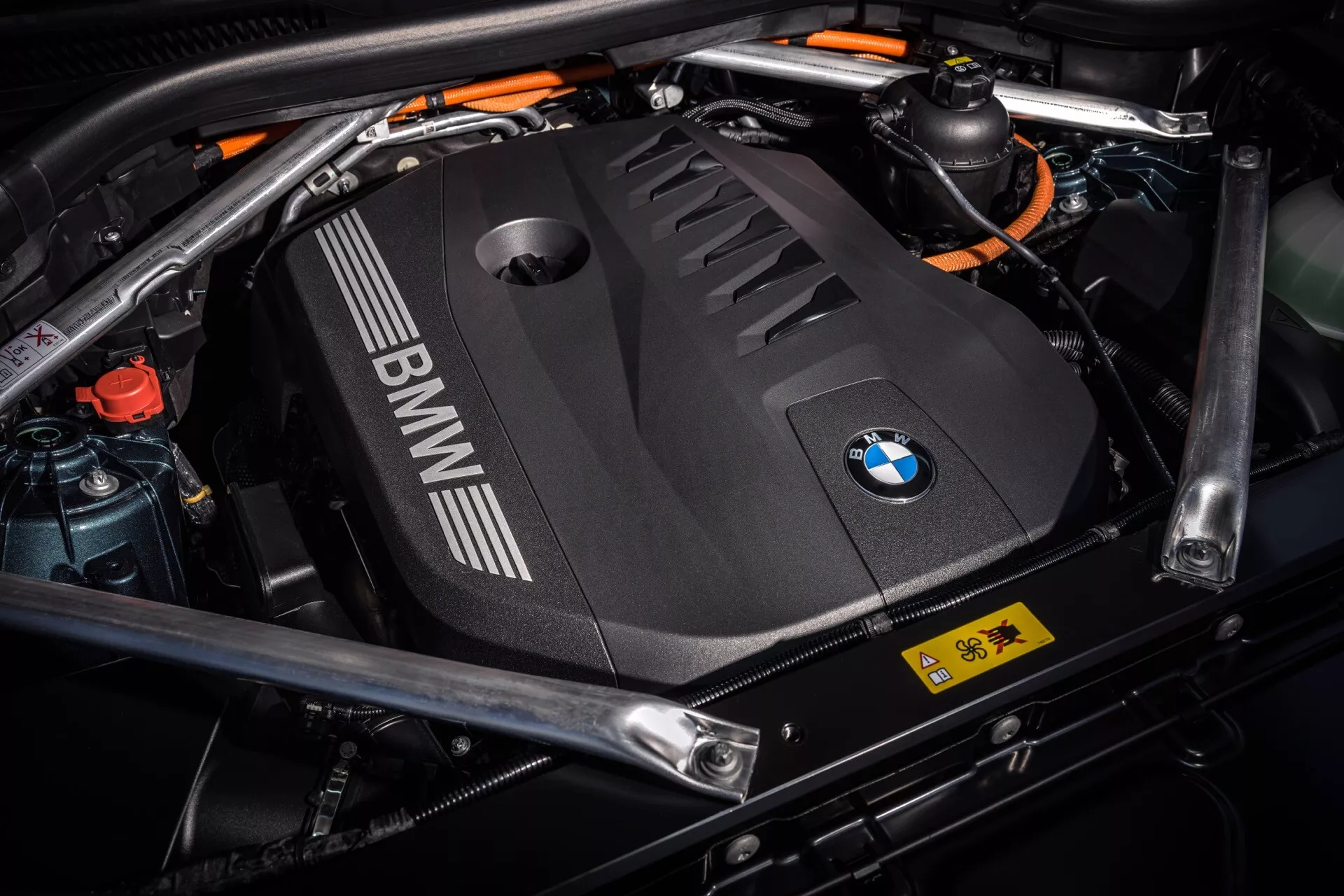 Närbild av motorutrymmet i BMW X5 2023 (G05-F95-Facelift) som visar det svarta motorlocket med BMW-logotyp, högspänningsorange kablar för en hybridmotor och säkerhetsetiketter, vilket betonar avancerad hybridteknologi och design detaljer.