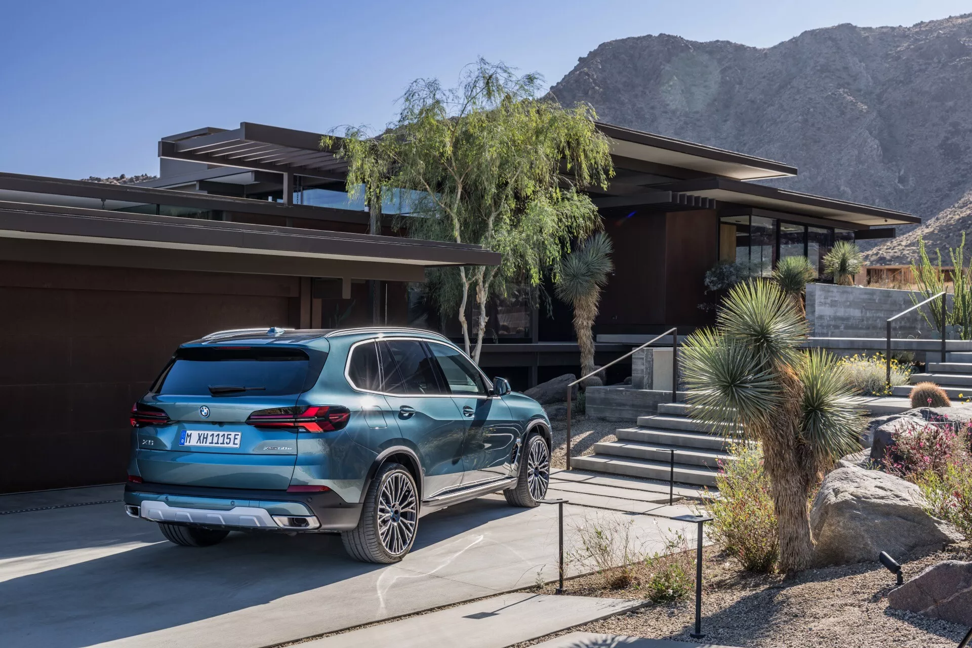 BMW X5 2023 (modell G05 F95 ansiktslyft) i medium blå färg parkerad utanför ett bostadshus i midcentury-modern stil, som visar bak- och profilvy, flerspakiga lättmetallhjul, parkeringssensorer bak och horisontella bakljus