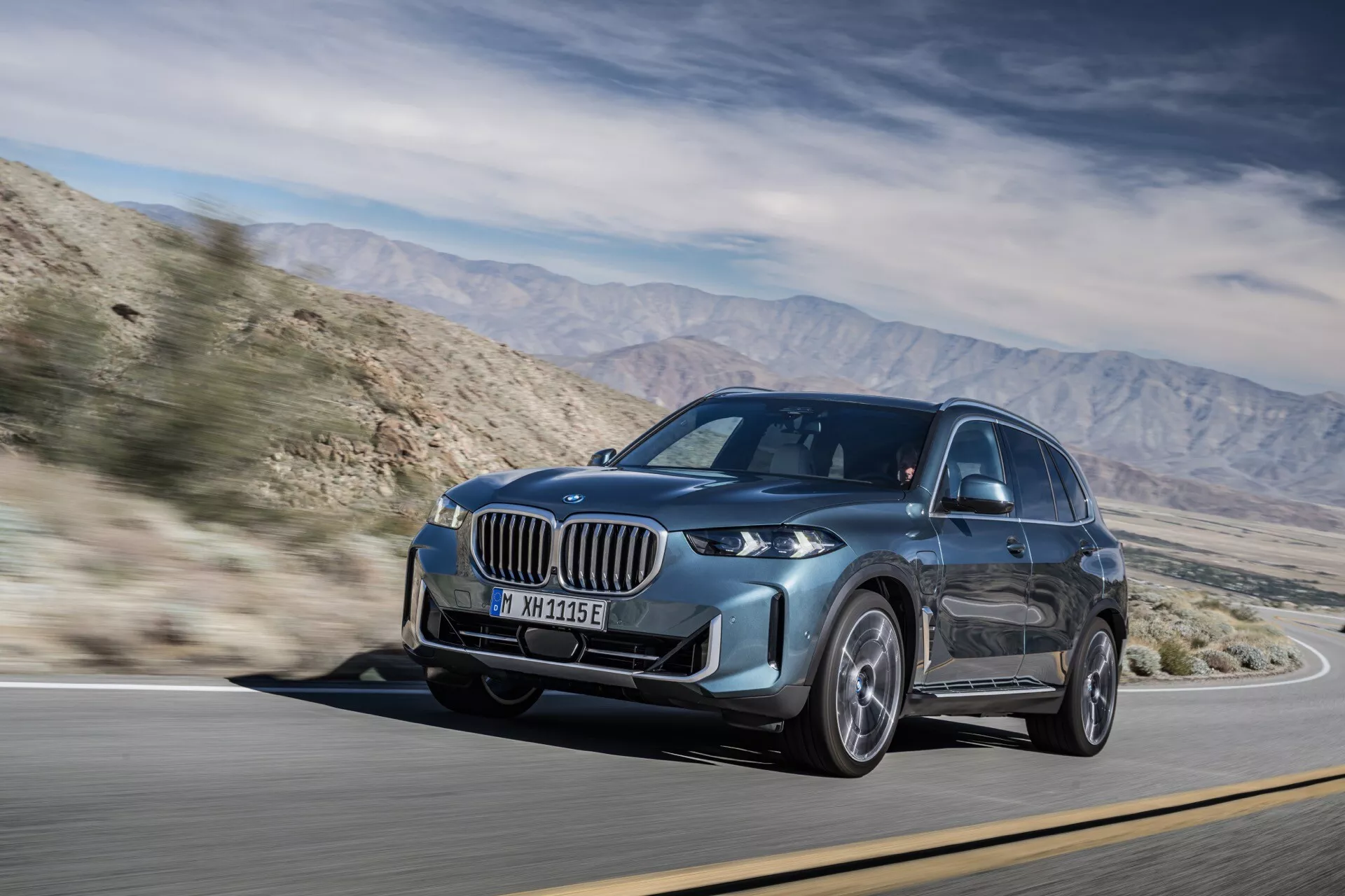 2023 BMW X5 (G05-F95) ansiktslyft modell i dynamisk rörelse, med en framträdande grill, LED-strålkastare och metallisk färg, fångad på en solig dag på en bergsväg