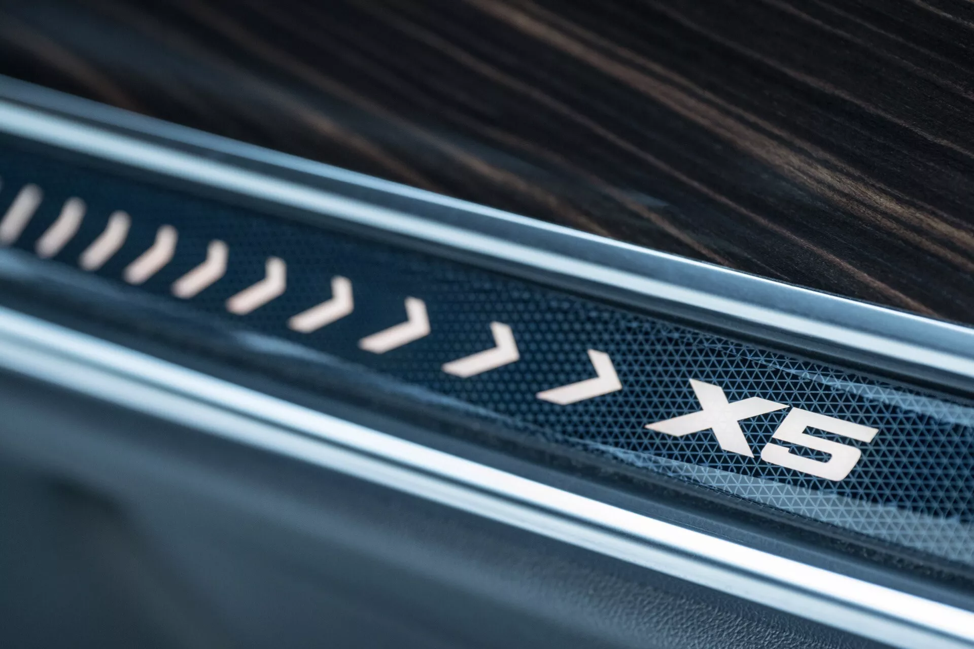 Närbild av interiören i BMW X5 2023 (G05-F95 facelift) som visar detaljer på den lyxiga tröskeln med 'X5' emblem, kolfibermönster och träfinish som betonar design och material av hög kvalitet